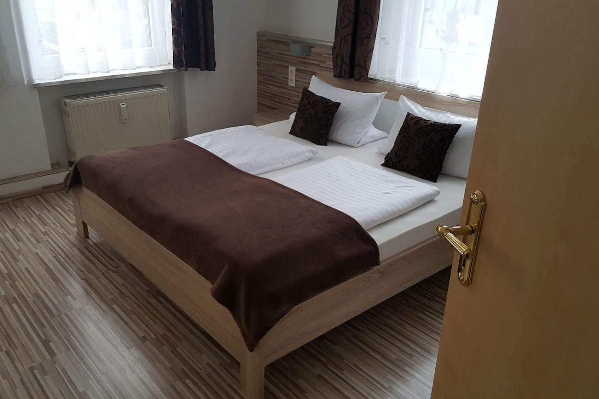 Un lit bien fait, recouvert d’une couverture marron, de draps blancs et agrémenté d’oreillers à motifs, dans une pièce au plancher en bois et dotée d’un radiateur.