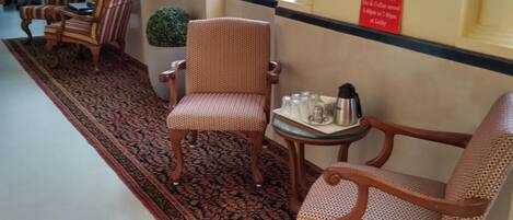 Un espacio de descanso con sillas estampadas y una mesa, un espejo decorativo, y un cartel que indica servicio de té y café de cortesía.