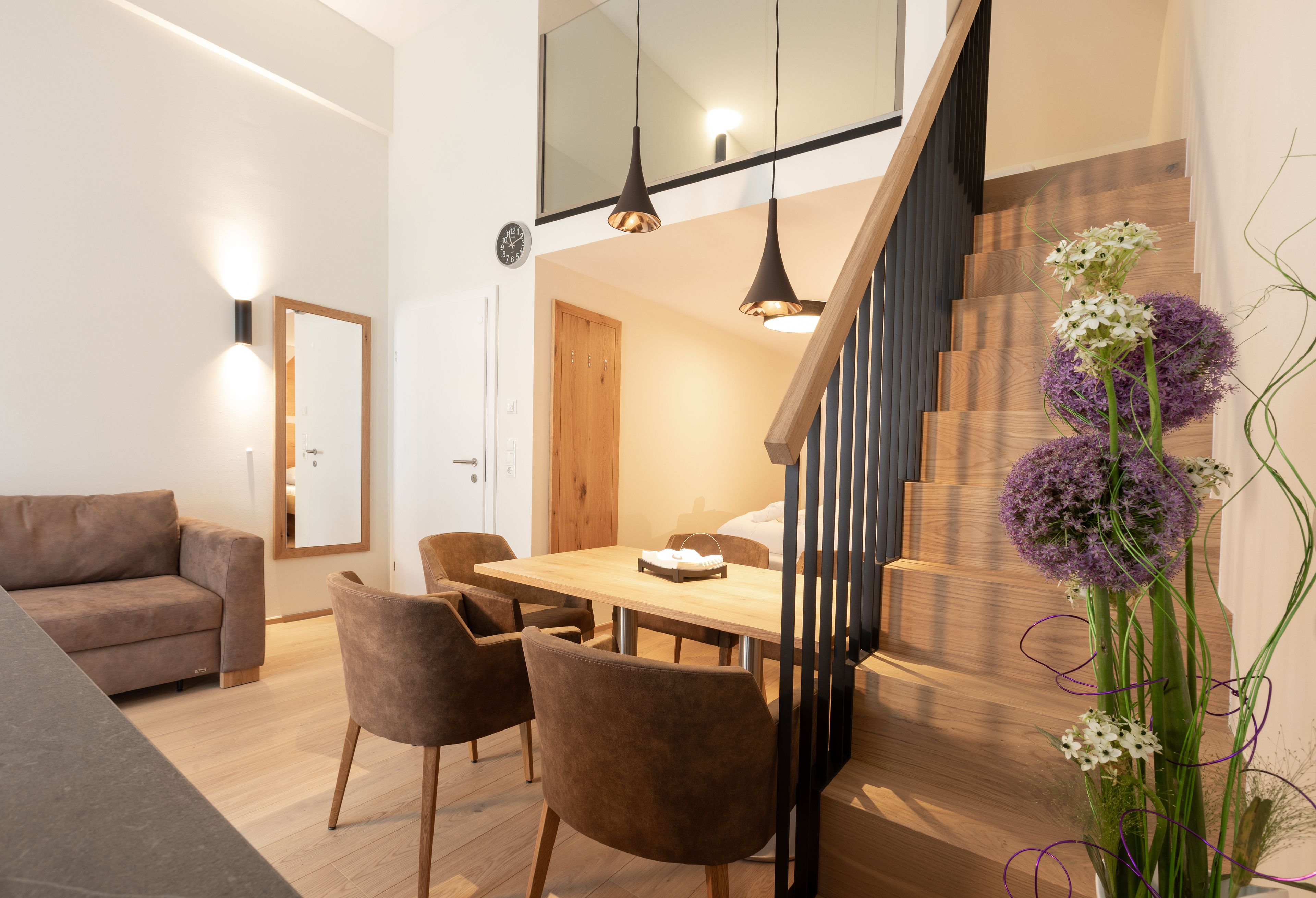 A modern living room with a wooden dining table, brown sofas, and a staircase with wooden steps.