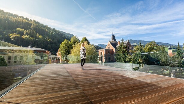 A wooden deck with a glass barrier, overlooking a mountainous landscape and a historic building with a pointed roof. - die Bergerin - Appartements Schladming (Schladming)