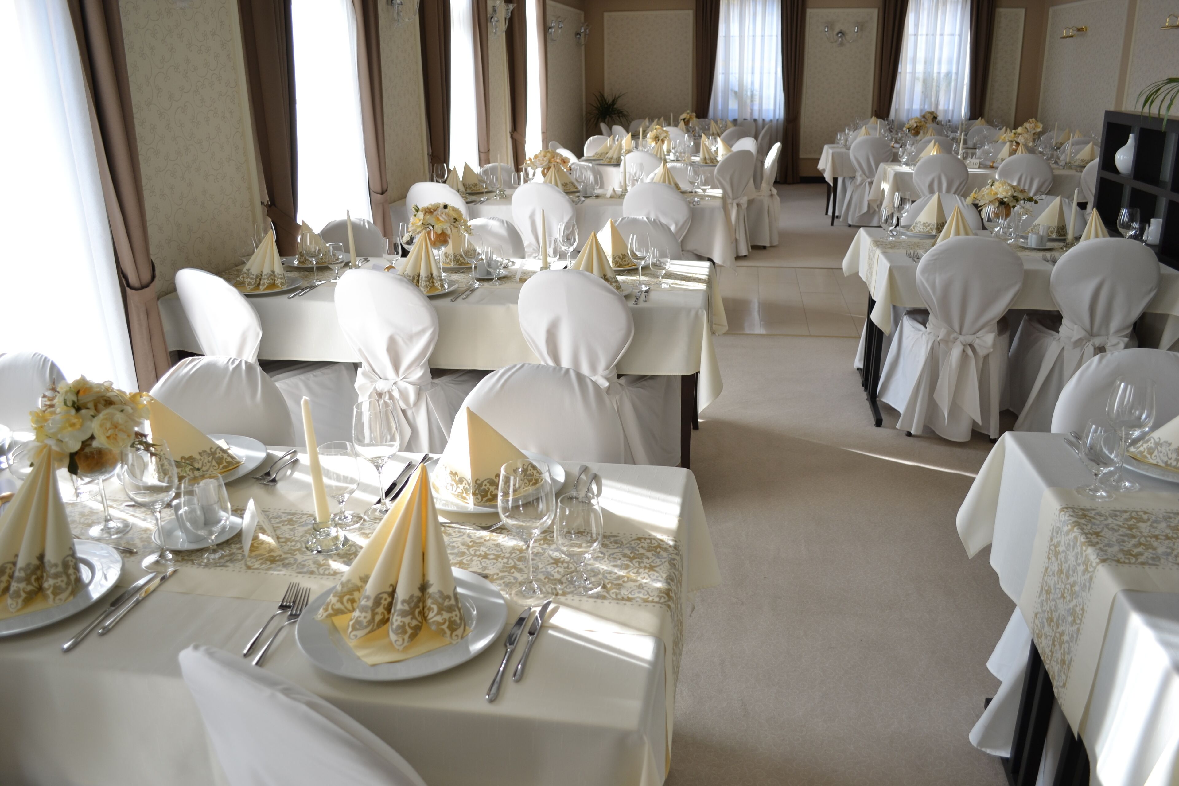 Un salón de banquetes con varias mesas dispuestas para un evento formal, destacando manteles blancos, sillas y centros de mesa con flores.