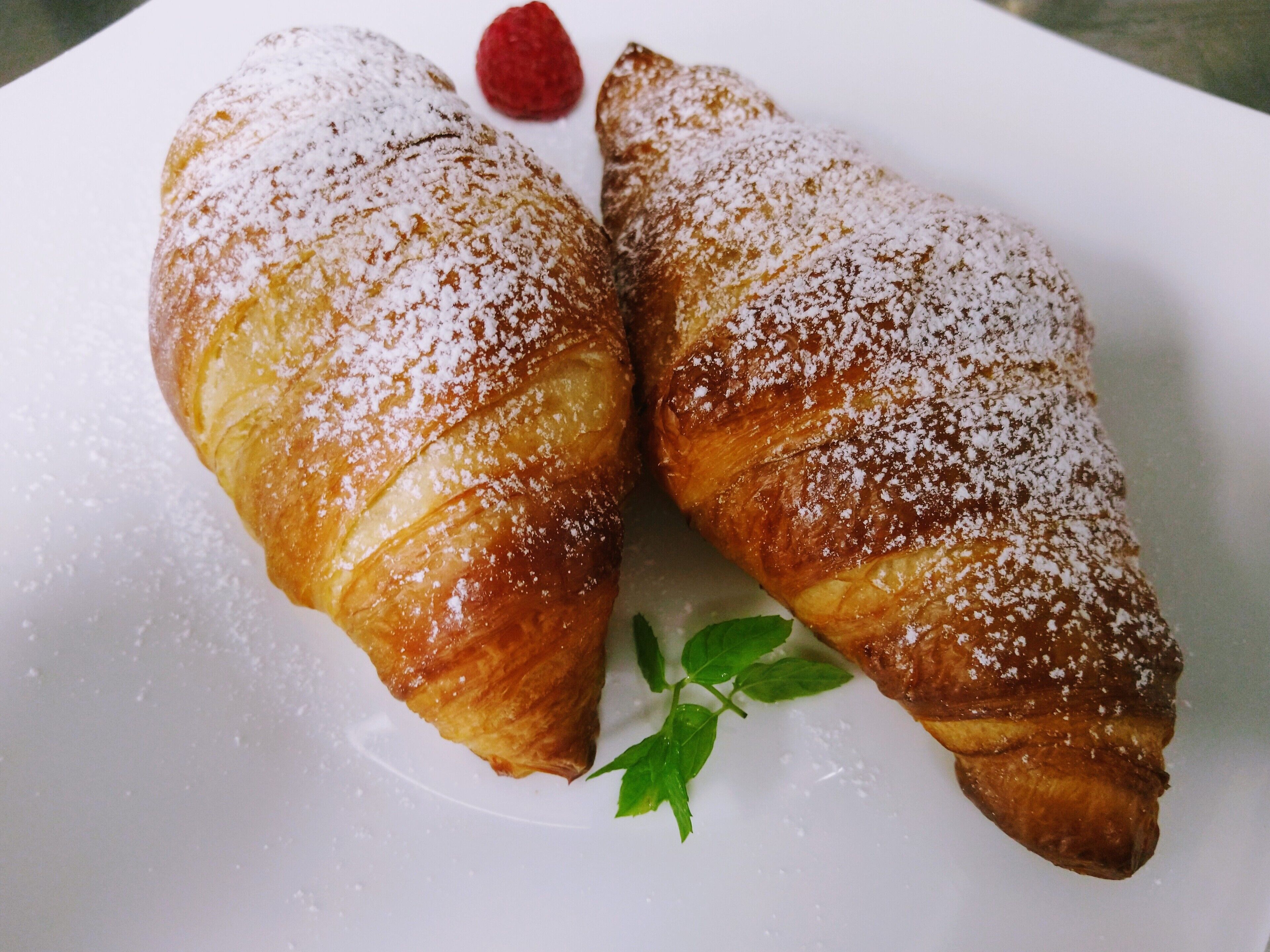 Twee croissants bestrooid met poedersuiker en een framboos op een wit bord.