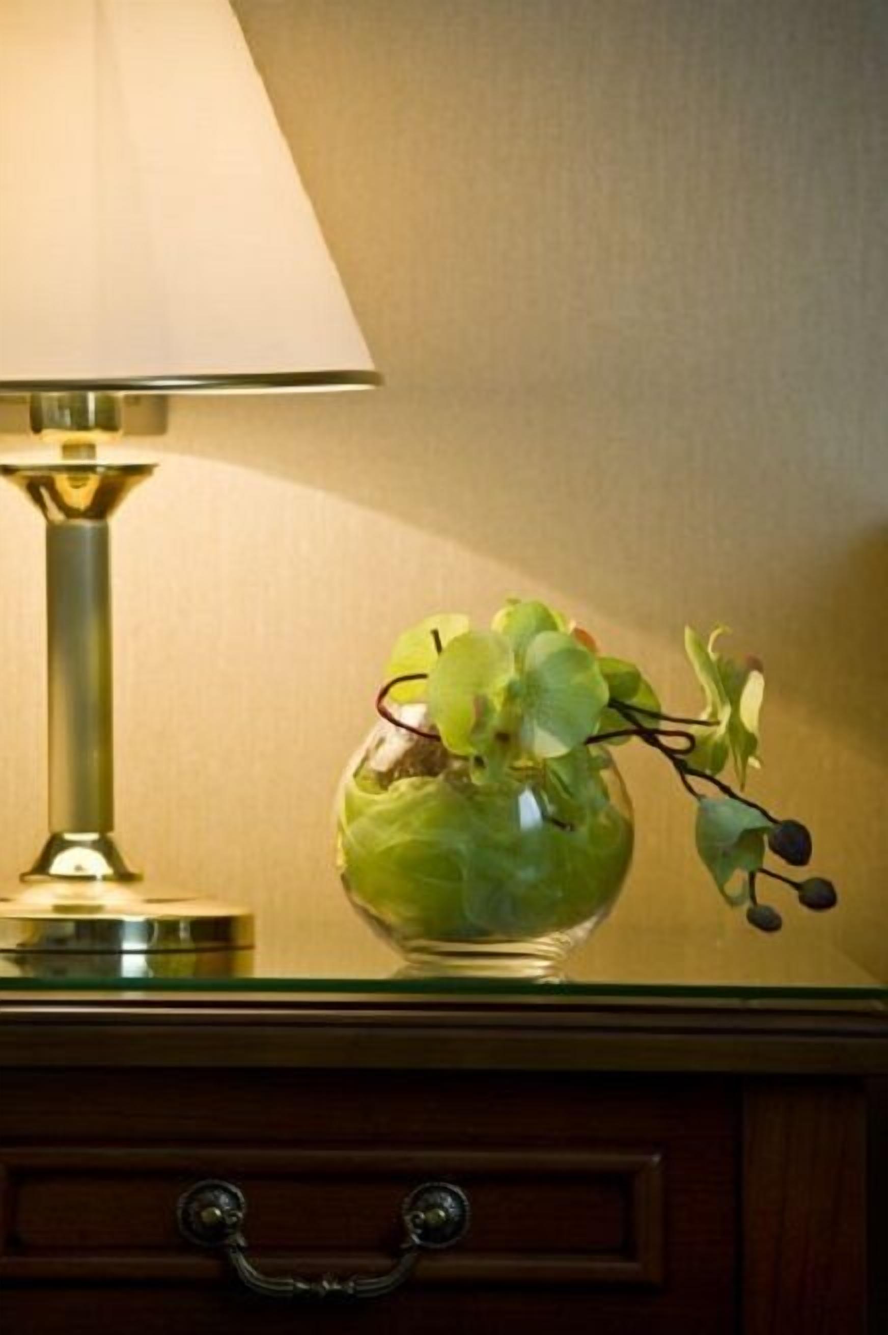 Una lámpara con pantalla color crema y un florero de vidrio con hojas verdes sobre una superficie de madera.