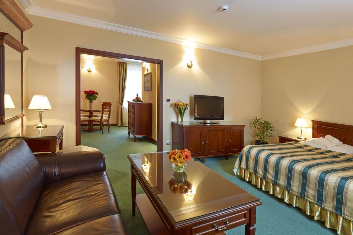 A hotel room with a leather sofa, a wooden coffee table with a vase of flowers, a bed with a striped bedspread, a television, and a dining area with a table and chairs.