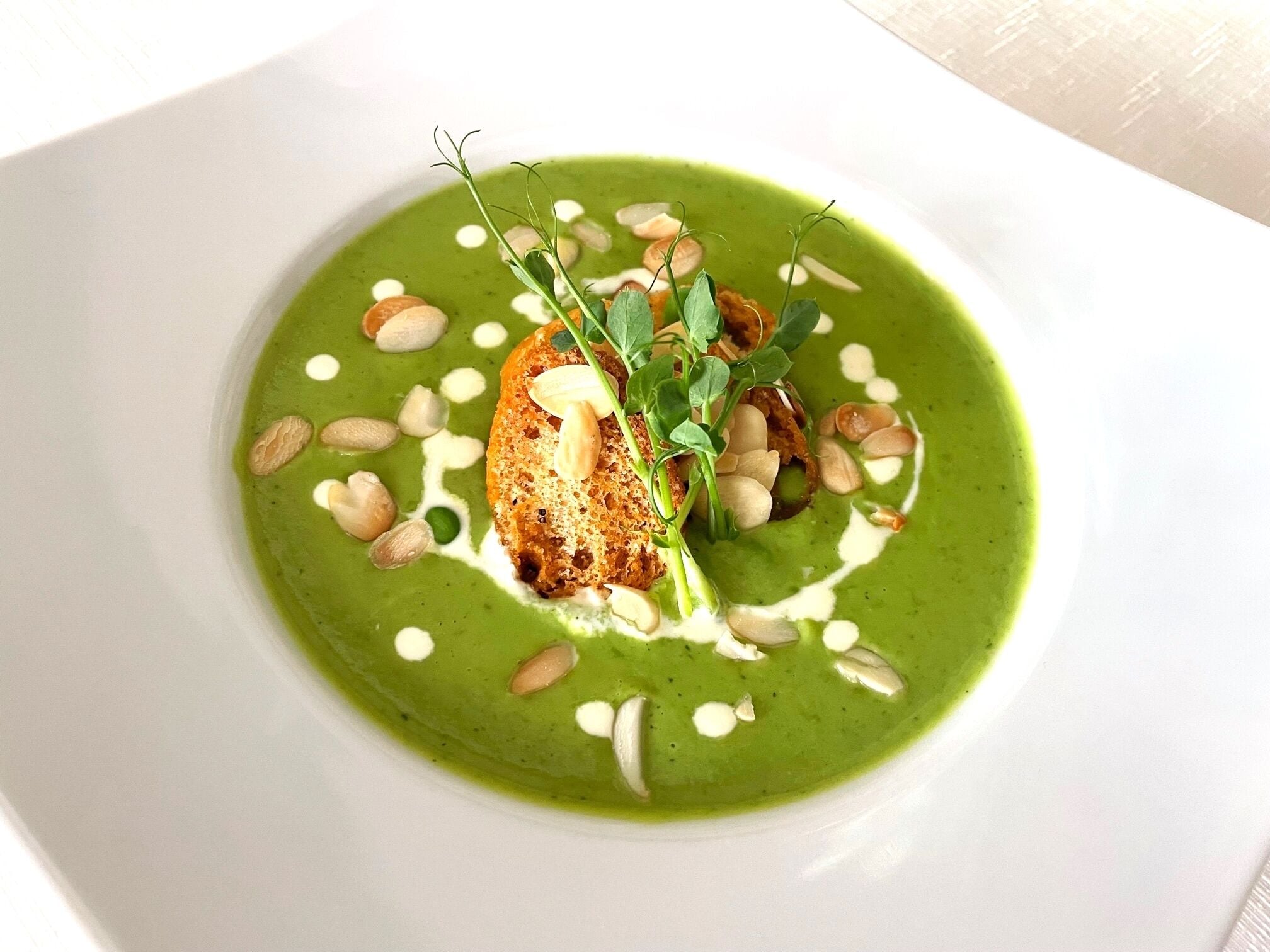 Une assiette blanche contenant une soupe verte, des amandes et un morceau de pain.
