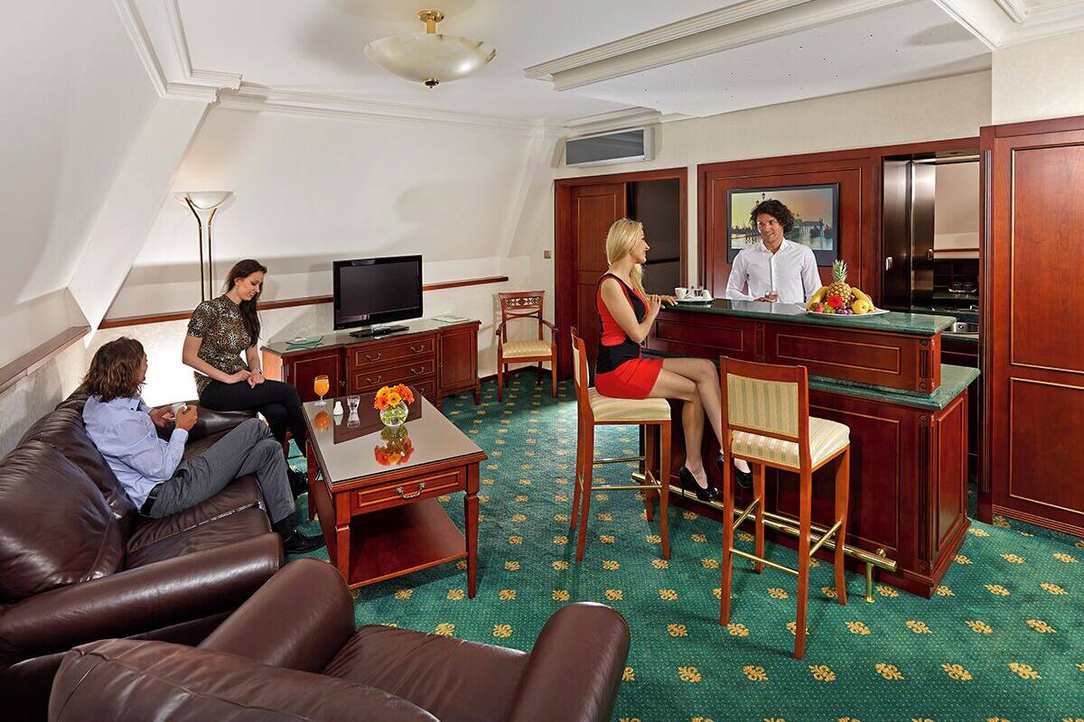 A hotel room with a living area, a kitchen, and a dining area. There are four people in the room, two on a couch and two at a table.