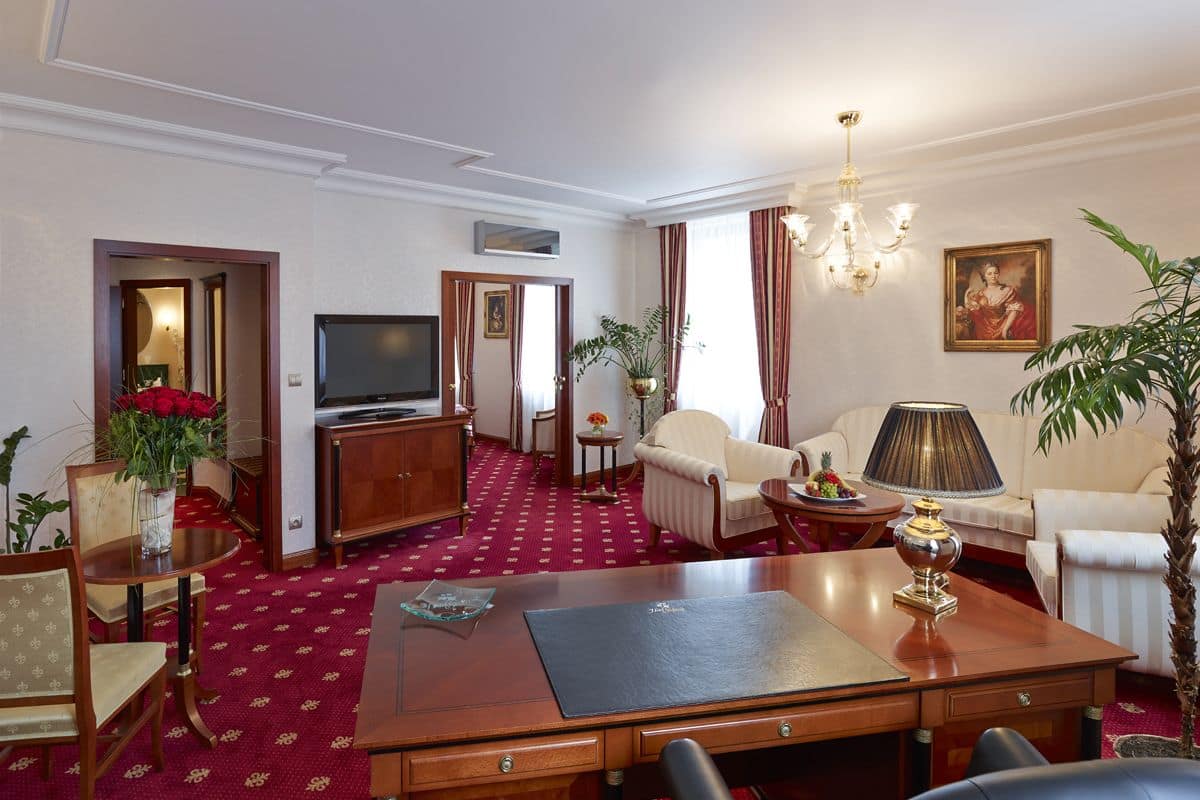 A hotel room with a large wooden desk, a television, a sofa, and a chair. There is a red carpet and a chandelier.