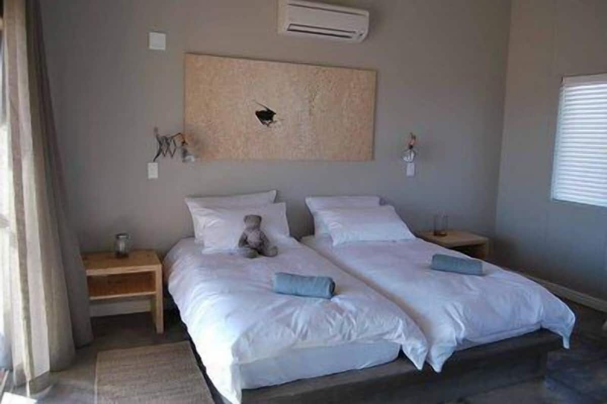 Una cama doble con ropa de cama blanca, un oso de peluche a la izquierda y una mesita de noche con una lámpara.
