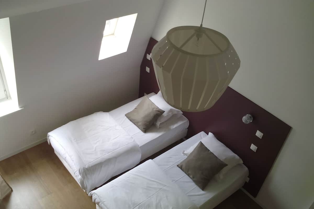A small, clean room with two single beds, a hanging light fixture, and a window providing natural light.
