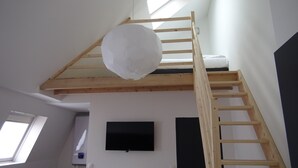A loft bedroom with a wooden ladder, a white hanging light fixture, and a flat-screen TV mounted on the wall. - Nationale AppartHotel (Lille)