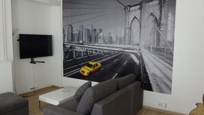 A living room with a large cityscape mural, a flat-screen TV, a grey sofa, and a white coffee table. - Nationale AppartHotel (Lille)