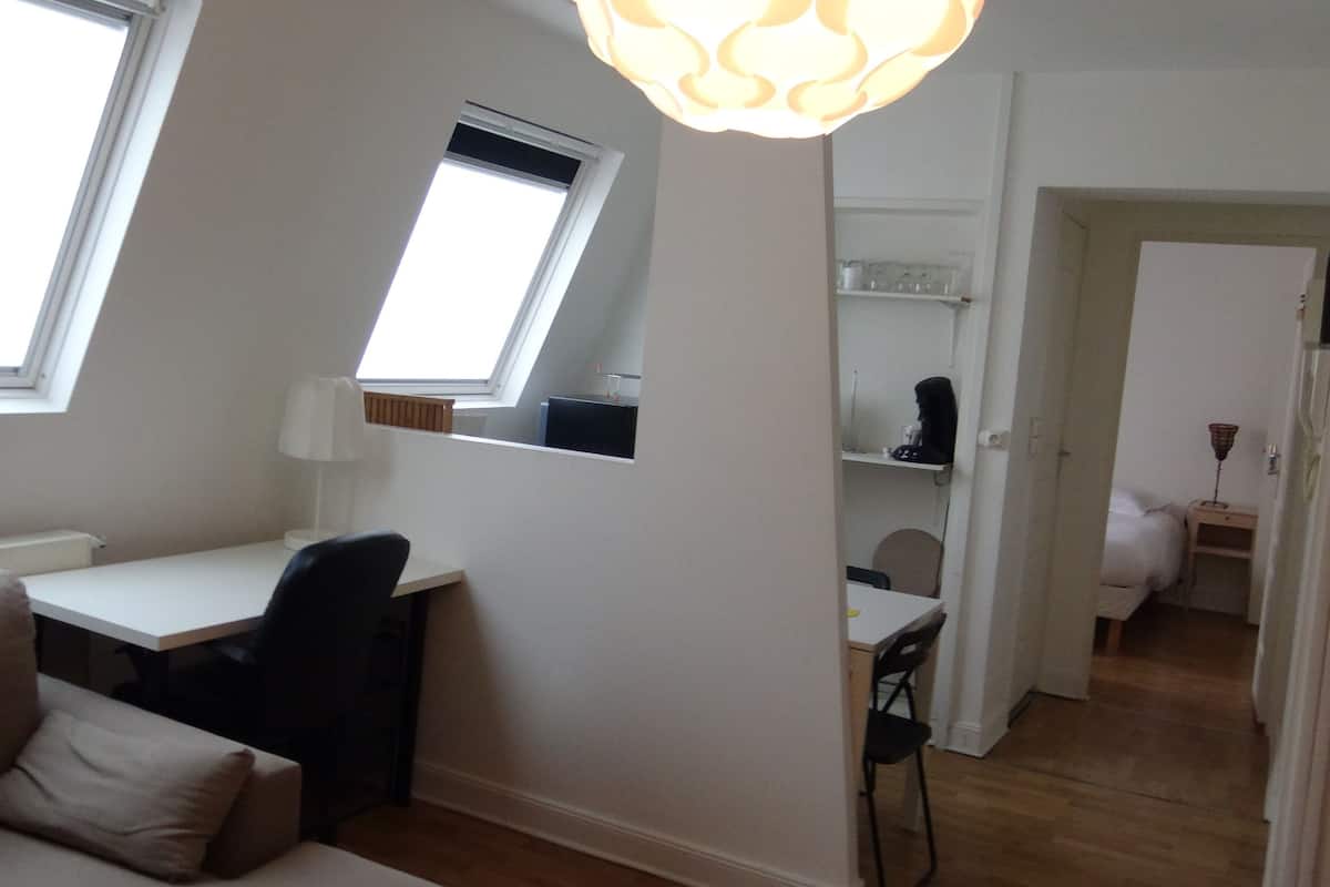 A small, well-lit room with a desk, chair, and a window. There is a ceiling light fixture and a door leading to another room.