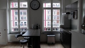 A kitchen with a dining table, chairs, a clock, and large windows with a view of buildings. - Nationale AppartHotel (Lille)