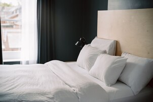 Una cama bien tendida con sábanas blancas y un cabecero de madera, una lámpara negra en la mesita de noche, y vistas de edificios a través de la ventana.