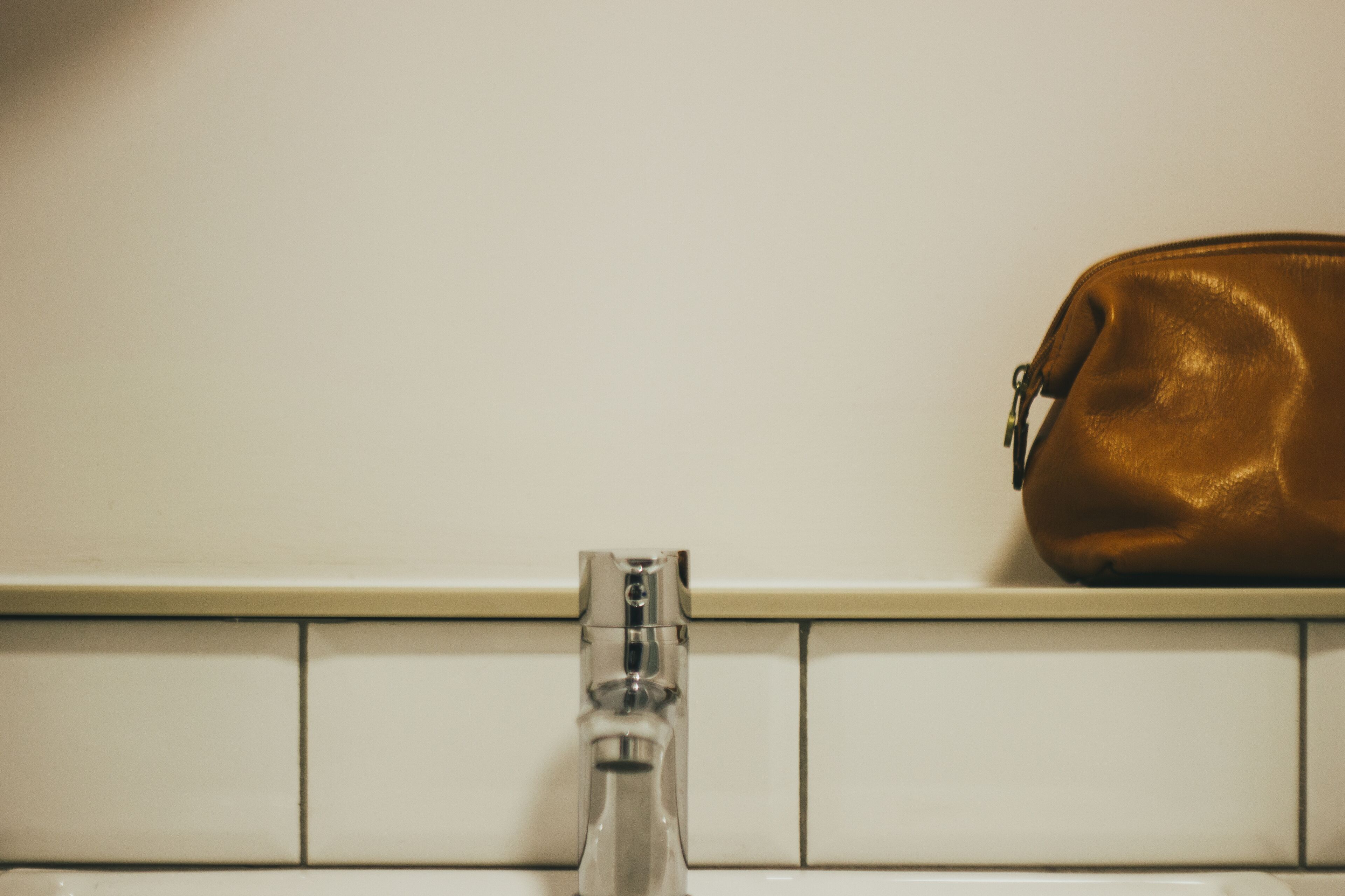 Un baño con pared de azulejos blancos, grifo cromado y bolso de cuero marrón en la pared.