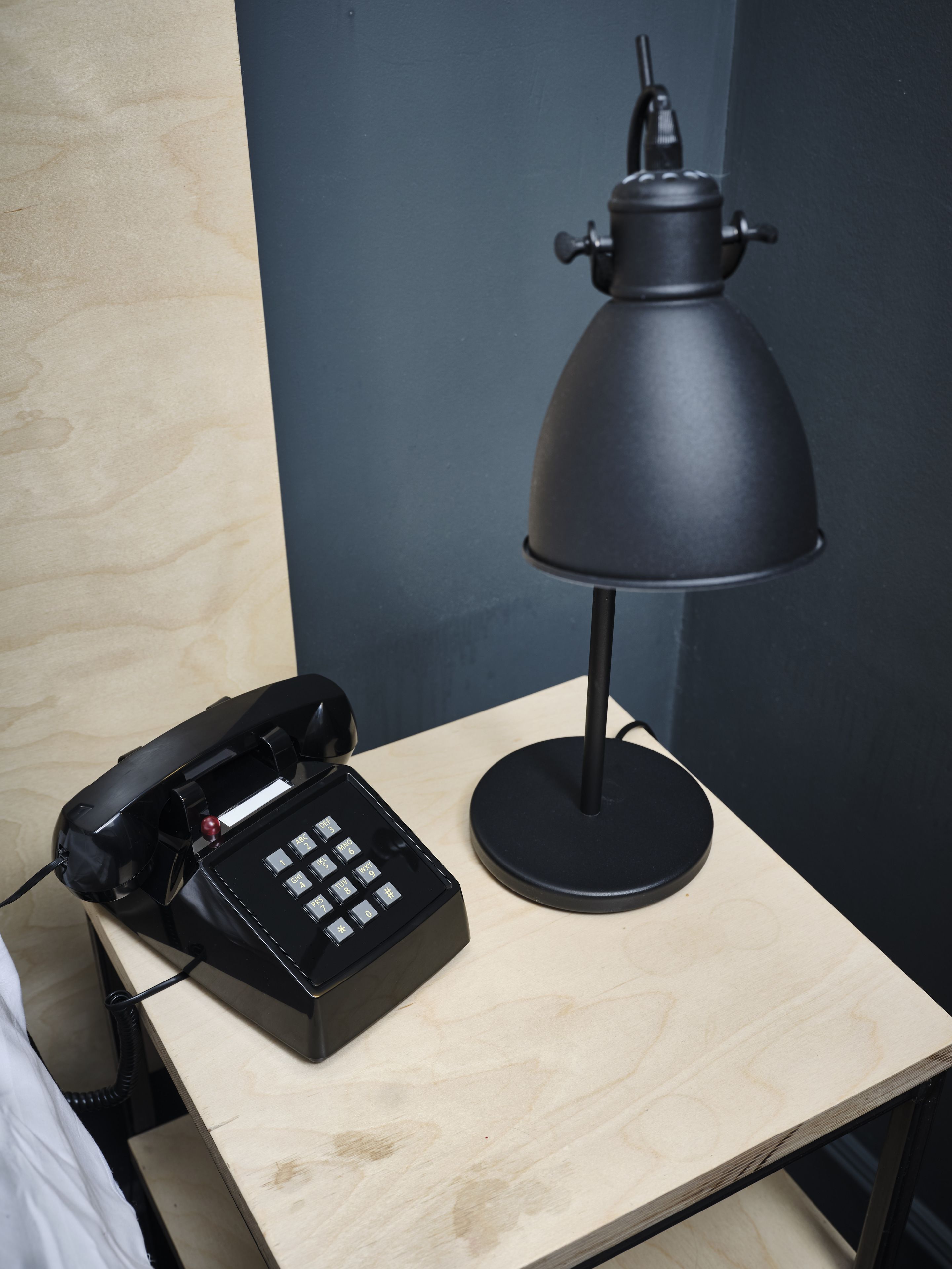 A wooden bedside table with a black telephone and a black desk lamp.