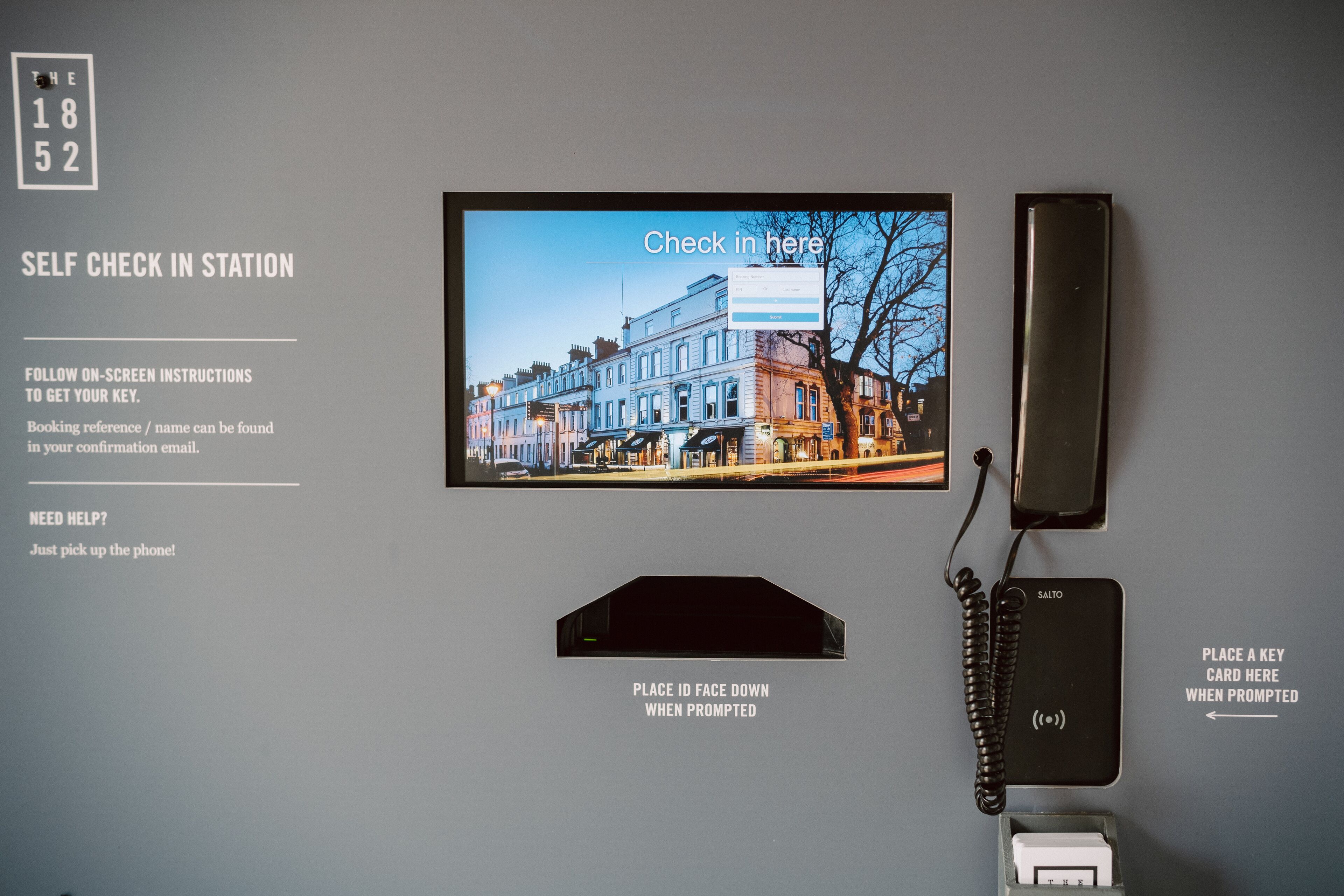 Una estación de auto-registro con un televisor mostrando un paisaje urbano, un teléfono y un teclado numérico. Se muestran instrucciones para el auto-registro.