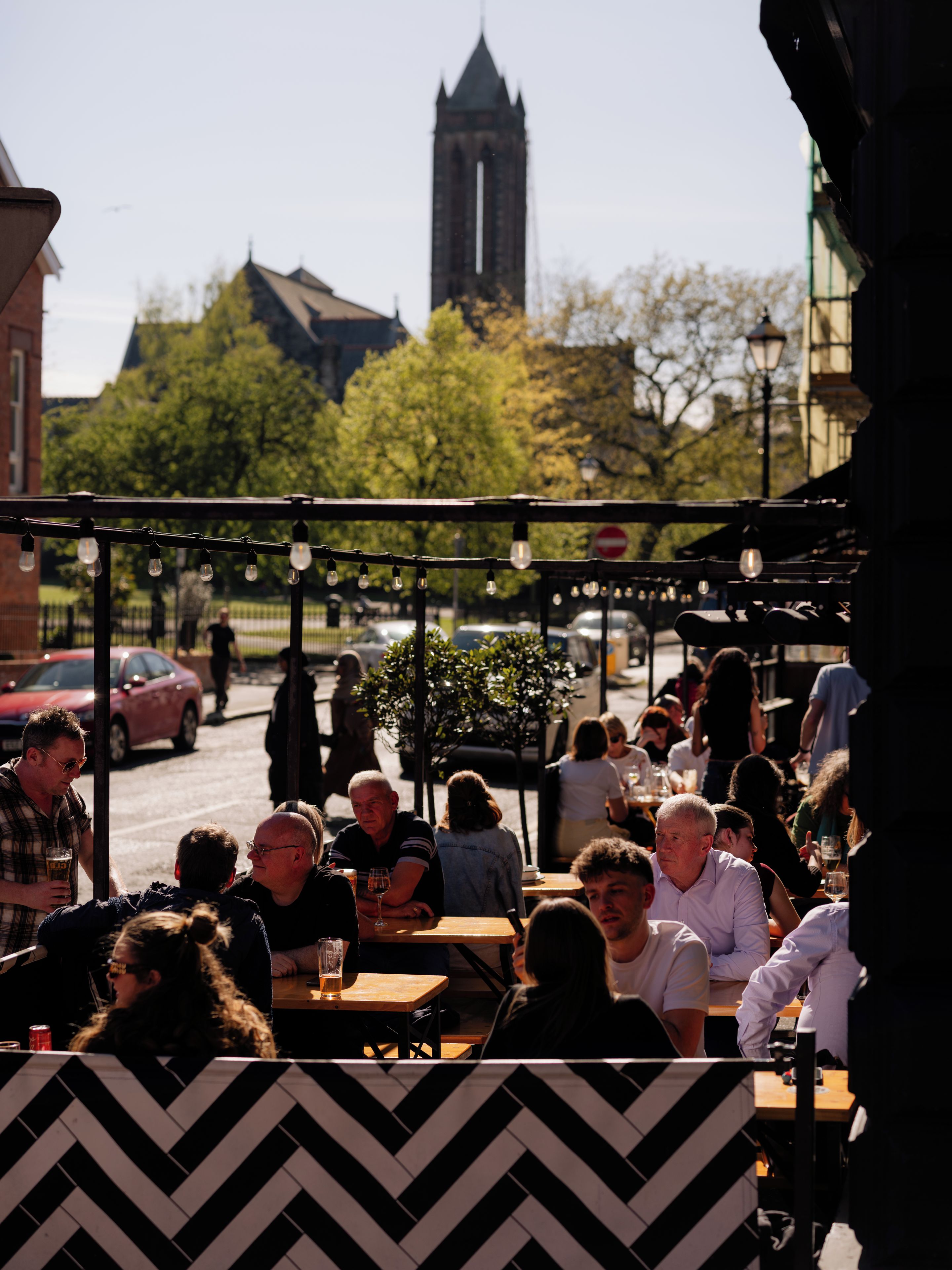 An outdoor seating area with tables and chairs, surrounded by trees and buildings, including a prominent tower in the background.