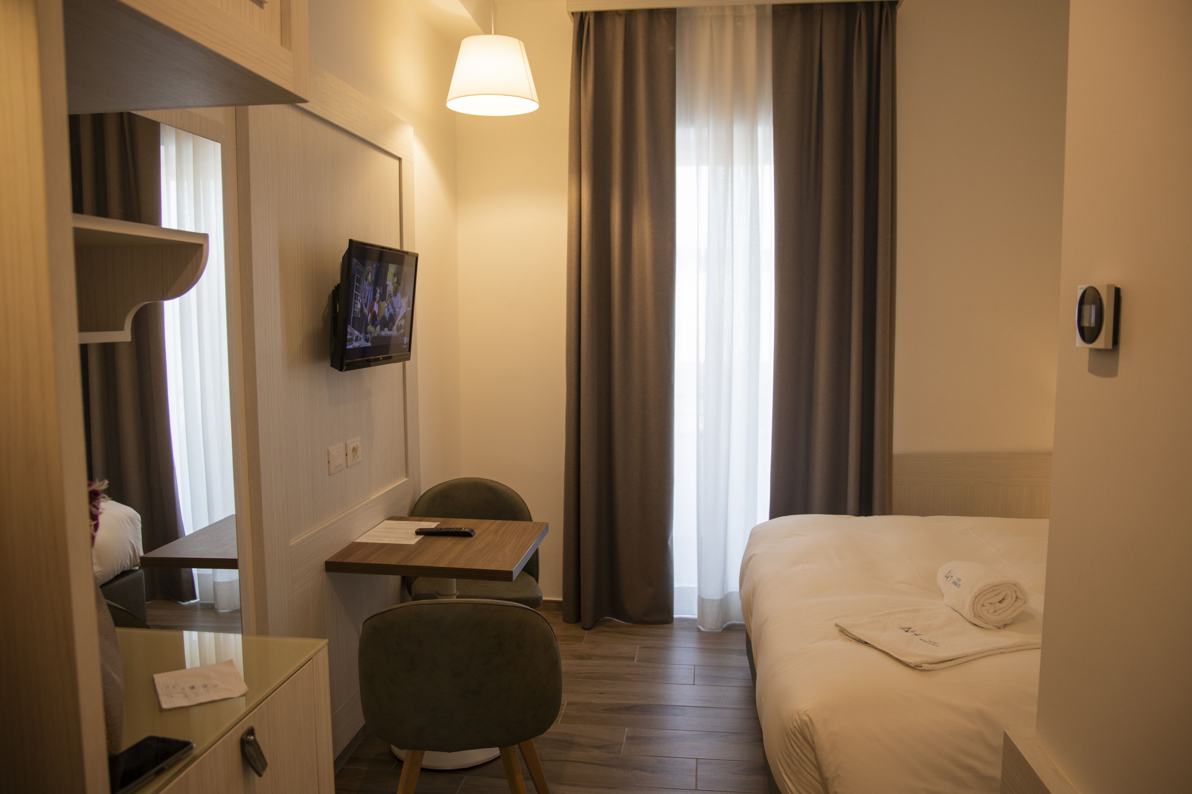 A hotel room with a bed, a desk with a chair, a TV mounted on the wall, and a window with curtains.