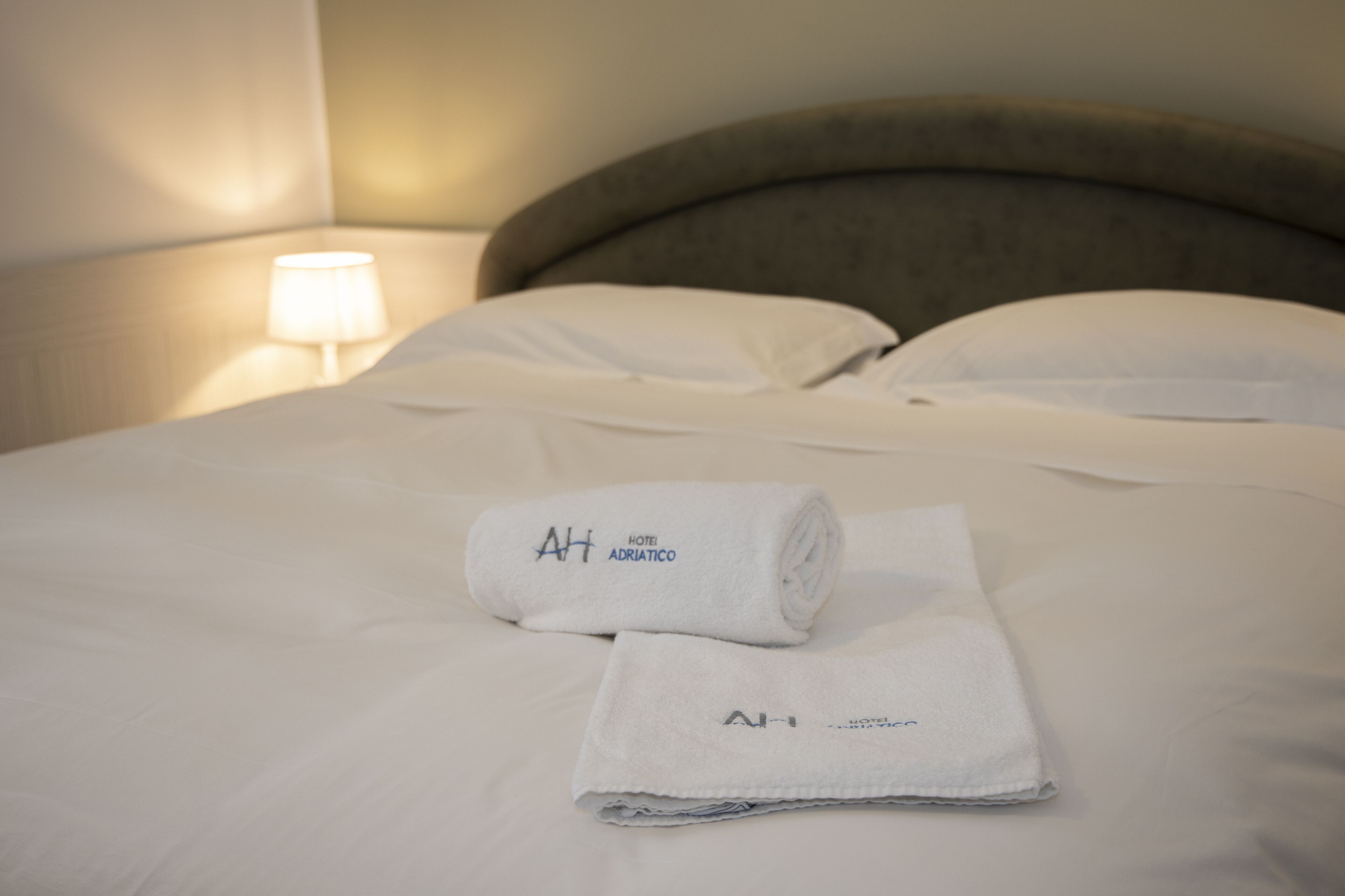 Un lit bien fait, avec du linge de lit blanc et deux serviettes enroulées, portant l’inscription « AH HOTEL ADRIATICO ».