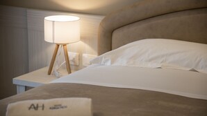 A neatly made bed with white linens and a beige headboard. A white lamp with a wooden base is on a bedside table. A rolled towel with "AH" is on the bed. - Hotel Adriatico (Tricase)