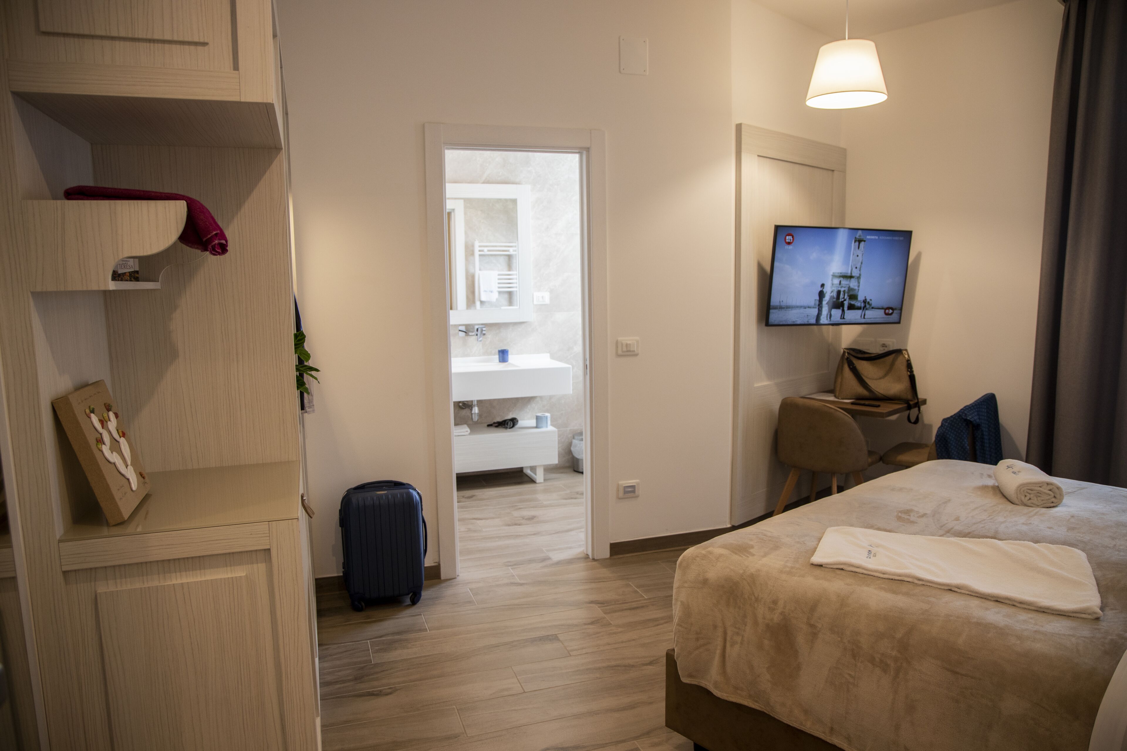 Une chambre d’hôtel avec un lit, un bureau, une télévision, une salle de bain visible à travers une porte ouverte et une valise posée sur le sol.
