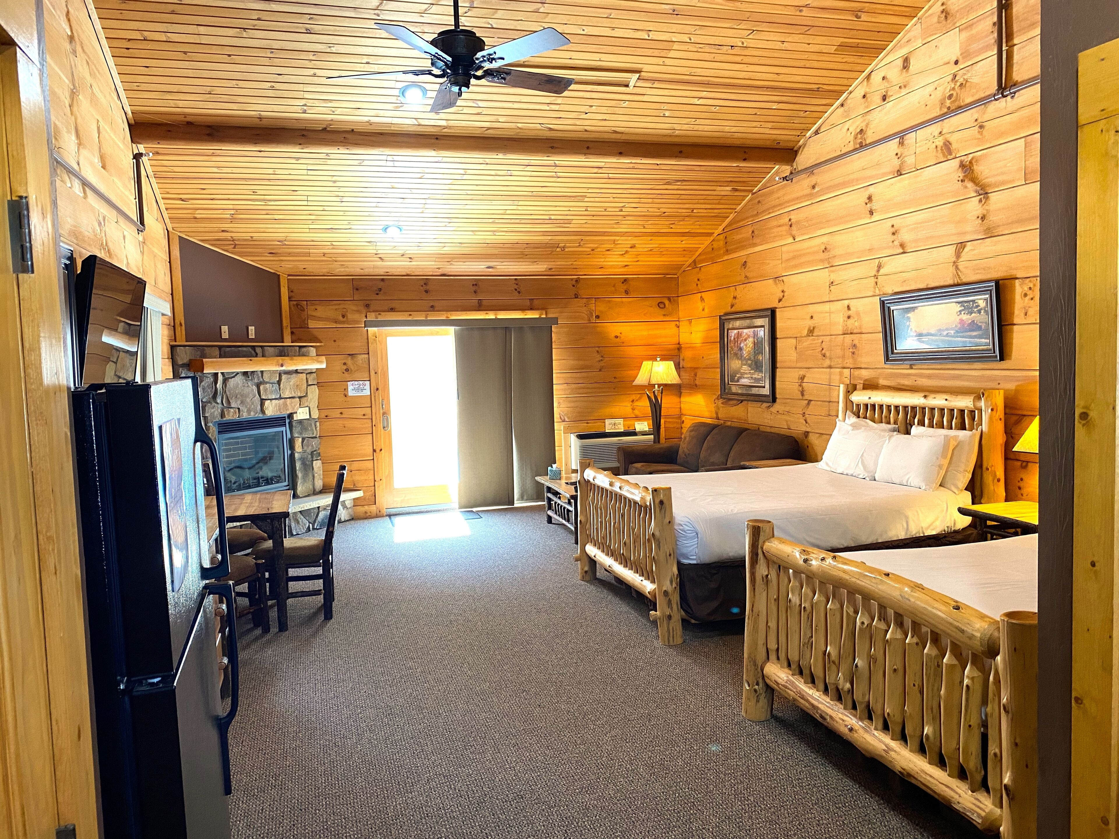 A log cabin room with a bed, a desk, a chair, a lamp, and a ceiling fan.