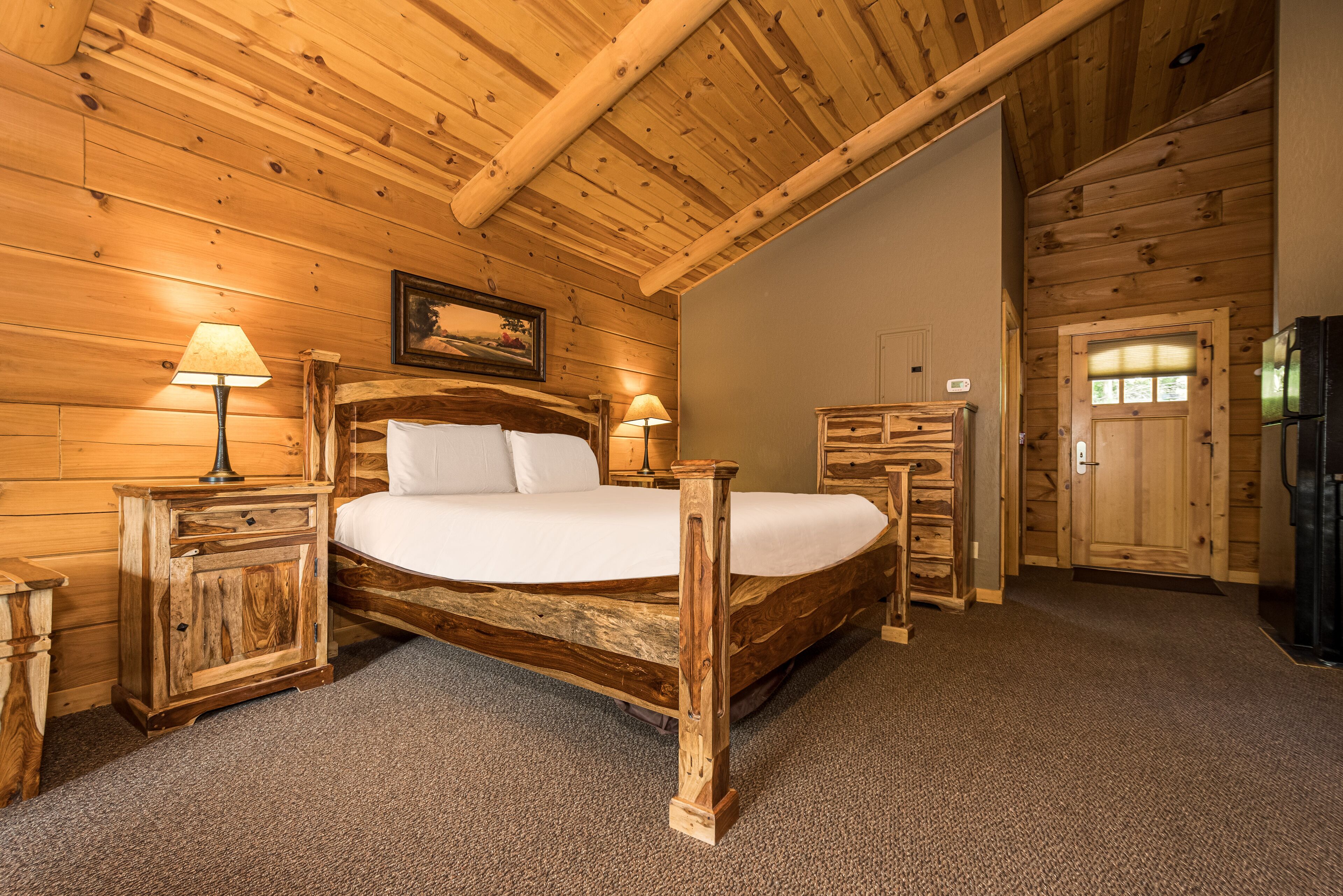 A wooden bedroom with a bed, nightstands, a dresser, and a door.