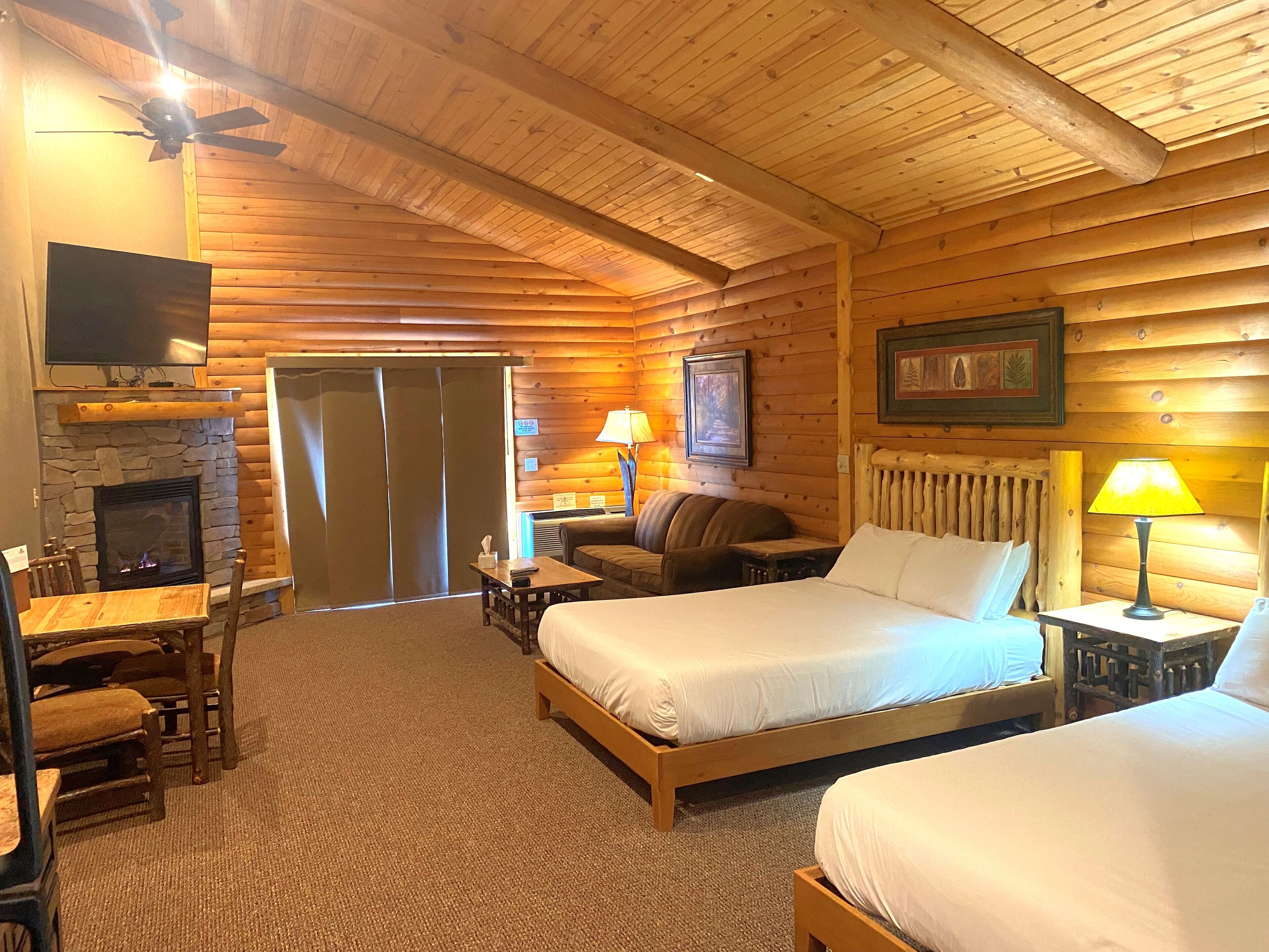 Ein Blockhauszimmer mit zwei Betten, einem Kamin aus Stein, einem Fernseher und einer Essecke mit Stühlen.