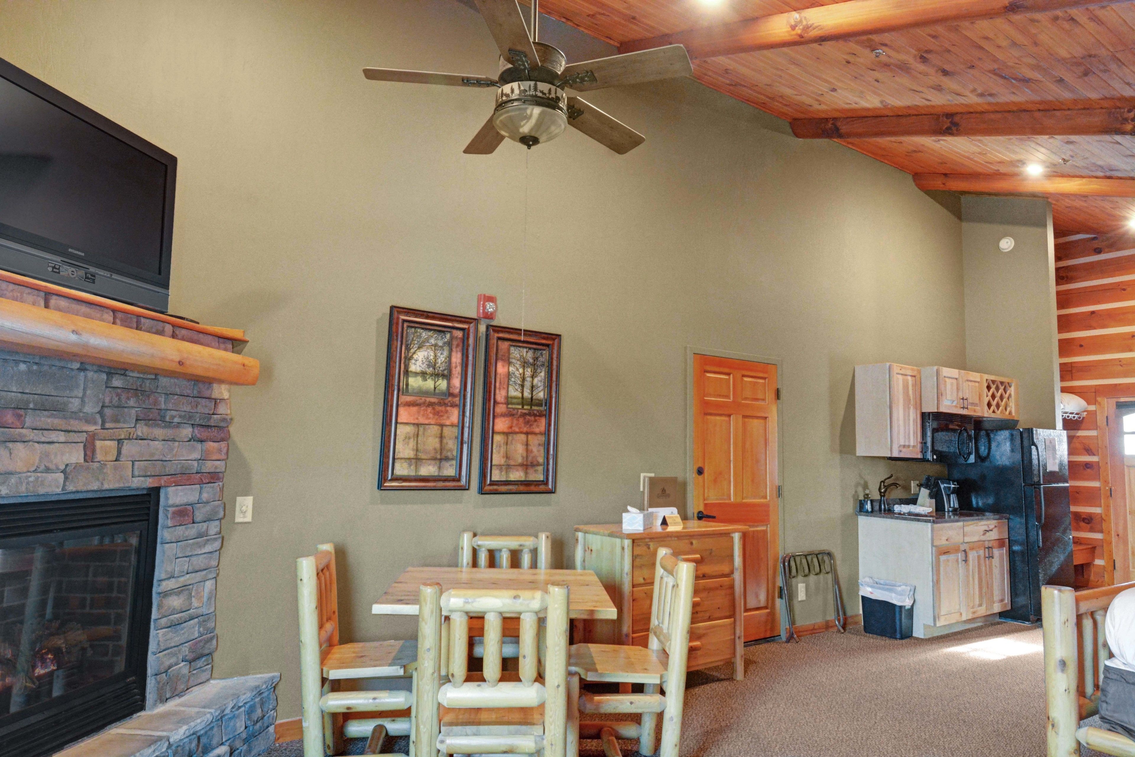 A cozy living area with a fireplace, wooden dining table, and chairs, a kitchen with cabinets, and a ceiling fan.