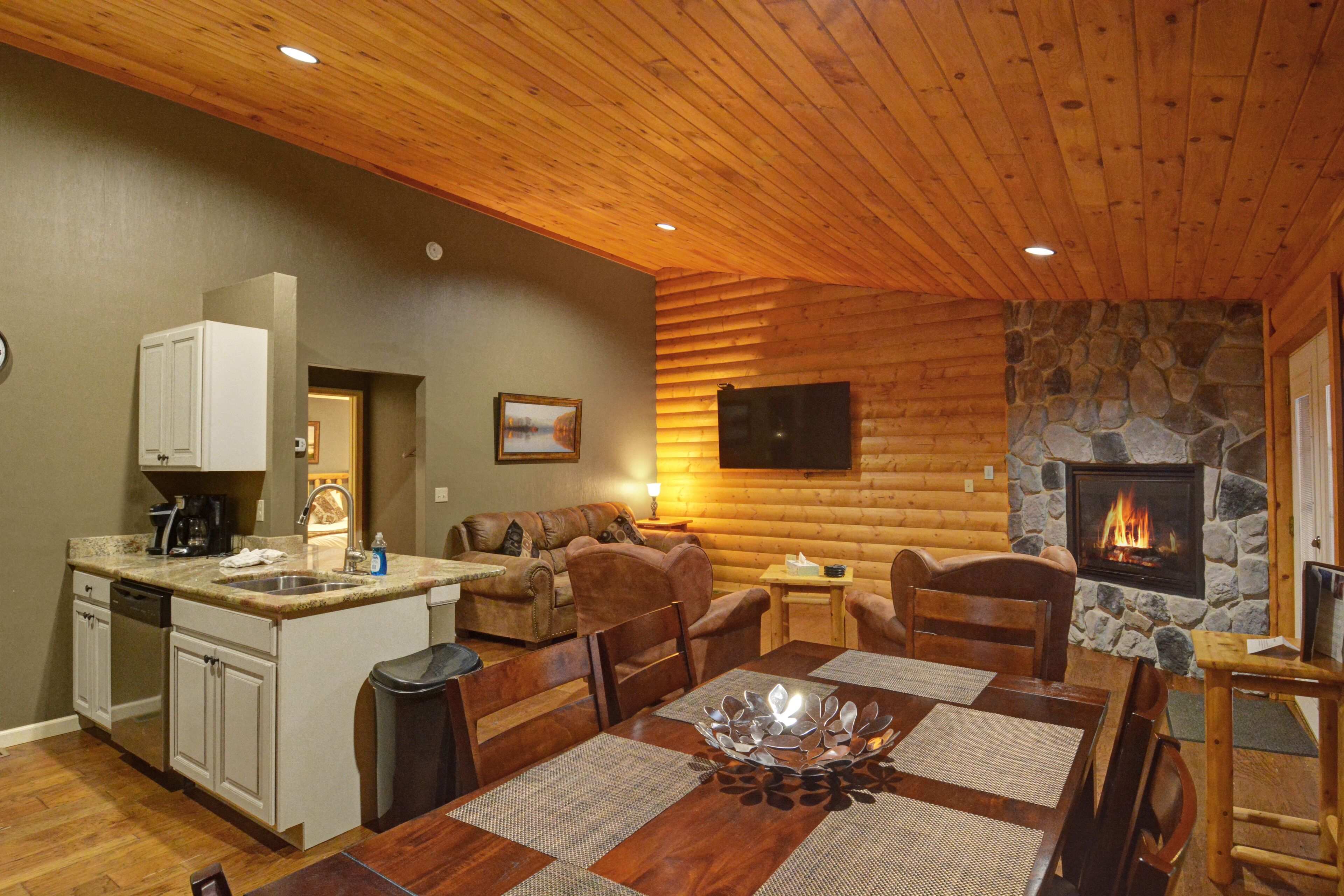 Acogedor interior de cabaña de troncos con chimenea de piedra, techo de madera y zona de comedor con una gran mesa de madera.
