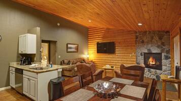 Acogedor interior de cabaña de troncos con chimenea de piedra, techo de madera y zona de comedor con una gran mesa de madera.