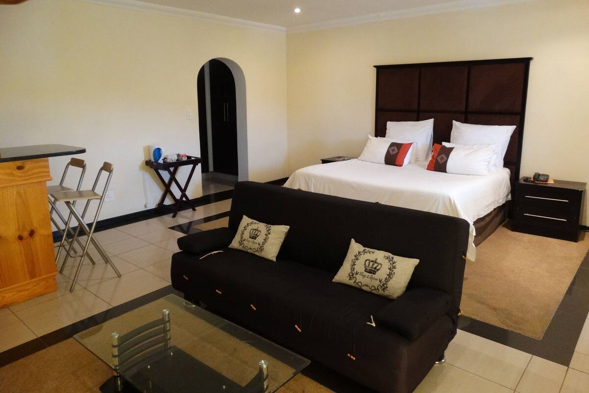 A hotel room with a bed, a black sofa, a glass coffee table, a wooden side table, and a small desk with chairs.