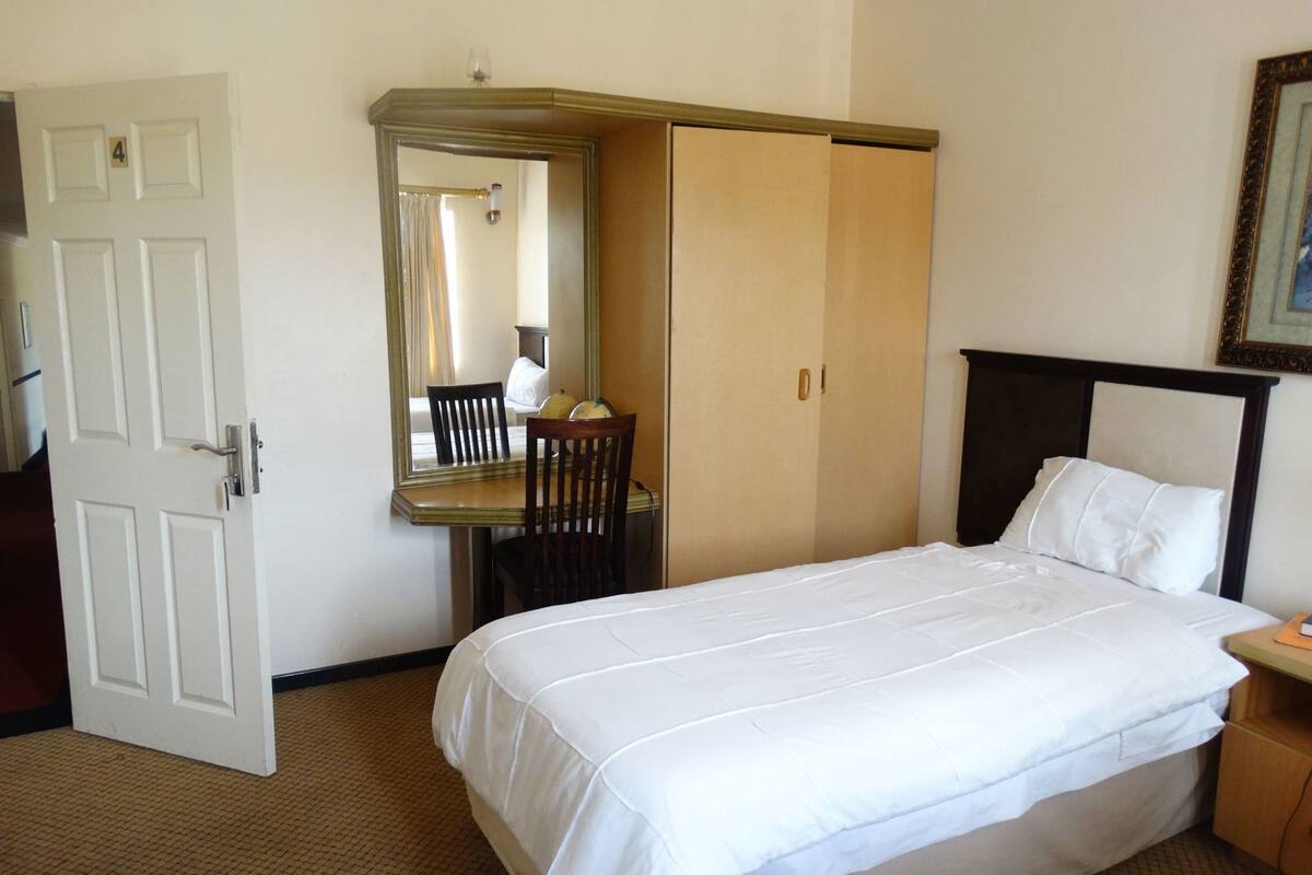 A hotel room with a bed, a desk with a chair, a wardrobe, and a mirror.