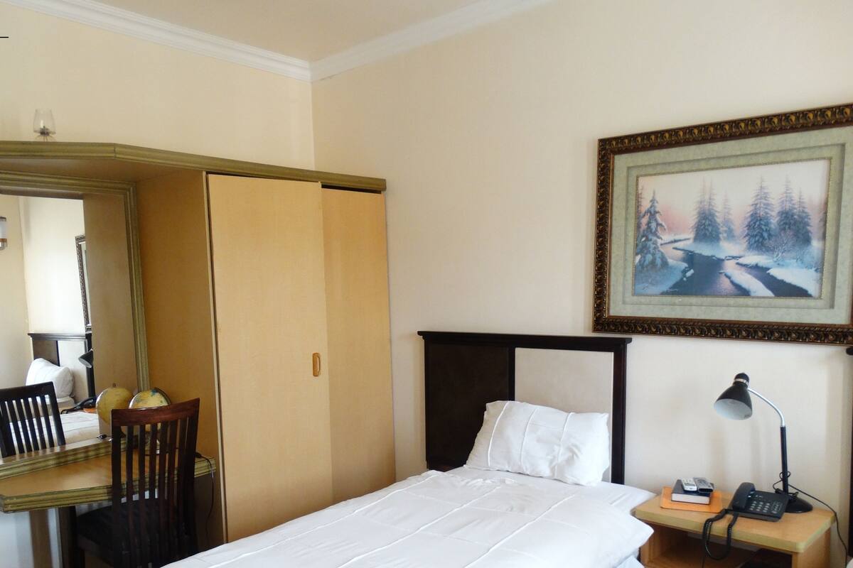 A hotel room with a bed, a desk with a chair, a nightstand with a lamp, and a painting on the wall.