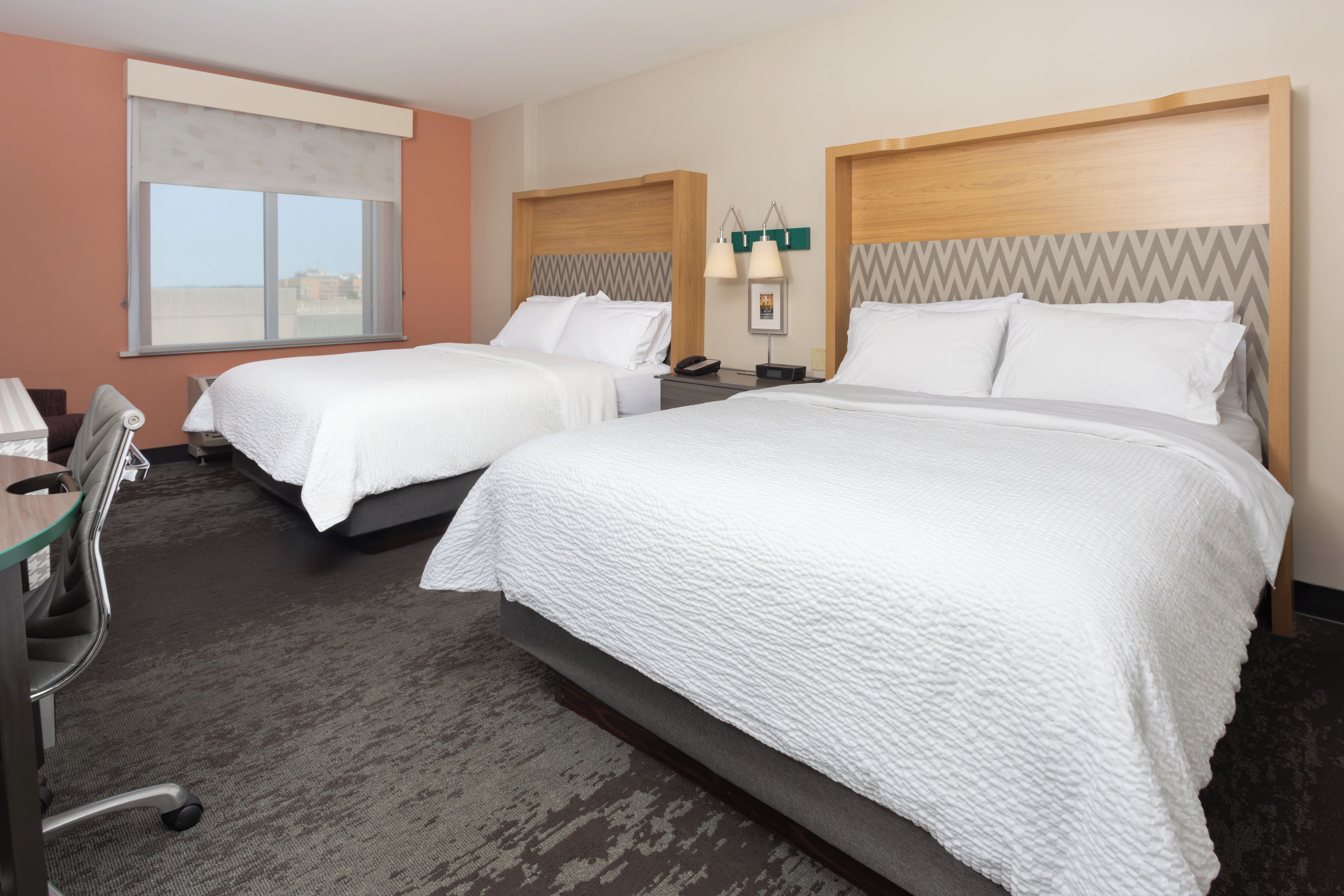 A hotel room with two beds, a desk, a chair, and a window.