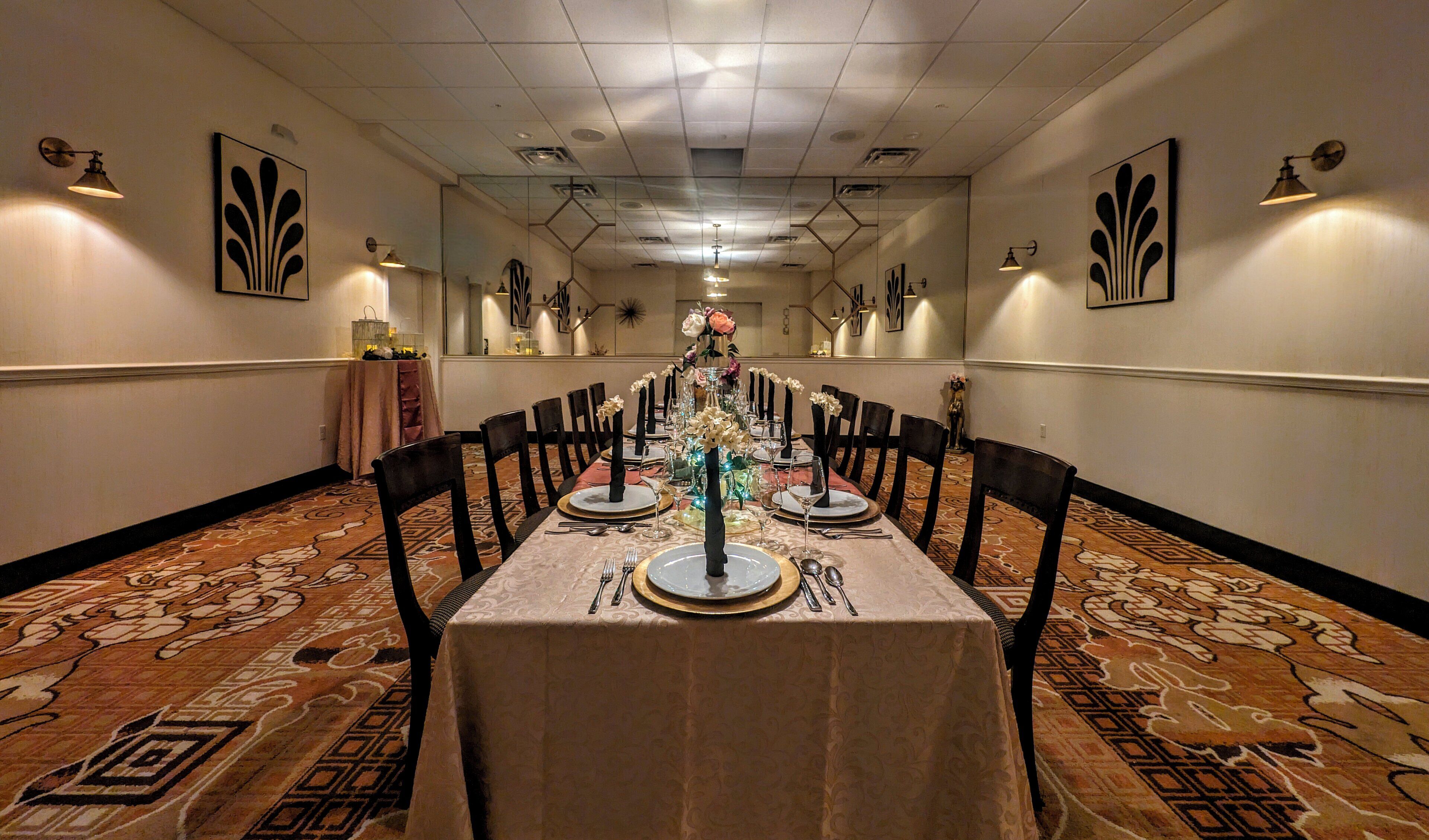 A long dining table set for a formal event, with chairs on both sides, in a room with patterned carpet and wall-mounted lighting.