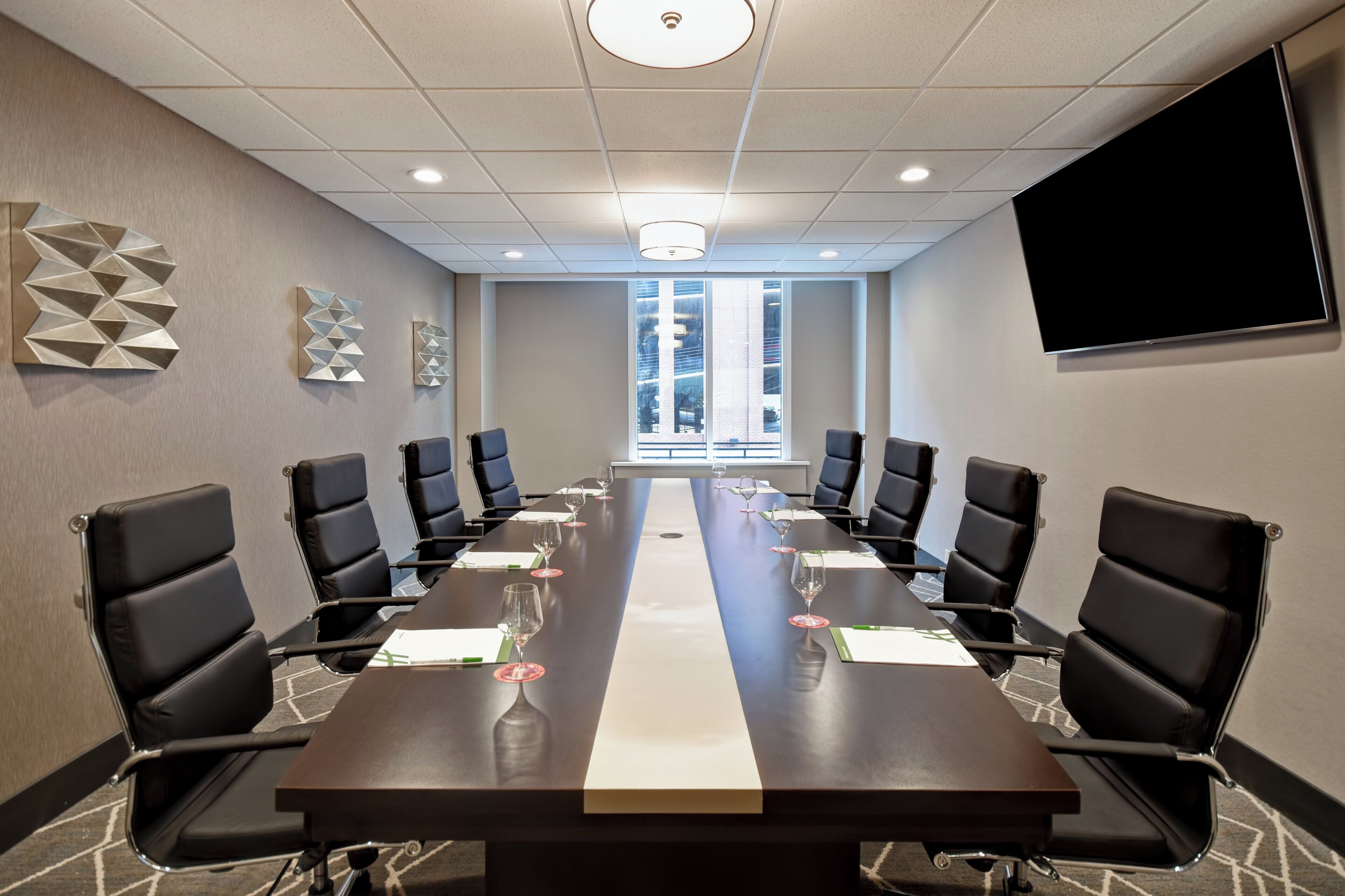 A conference room with a long table, black chairs, and a large screen on the wall.