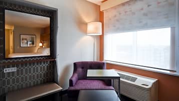 A hotel room with a purple armchair, a small table, a bench, a large window with a view, and a wall-mounted mirror.