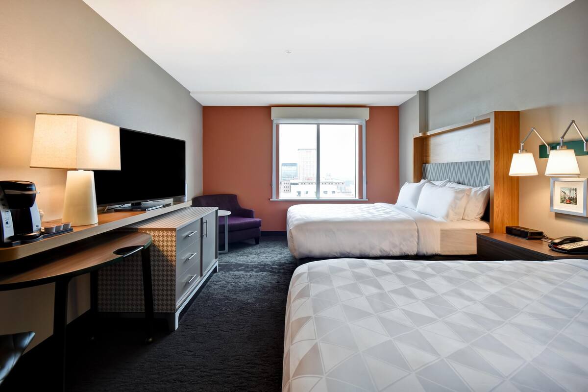 A hotel room with a bed, a desk with a TV, a chair, a window, and a lamp.