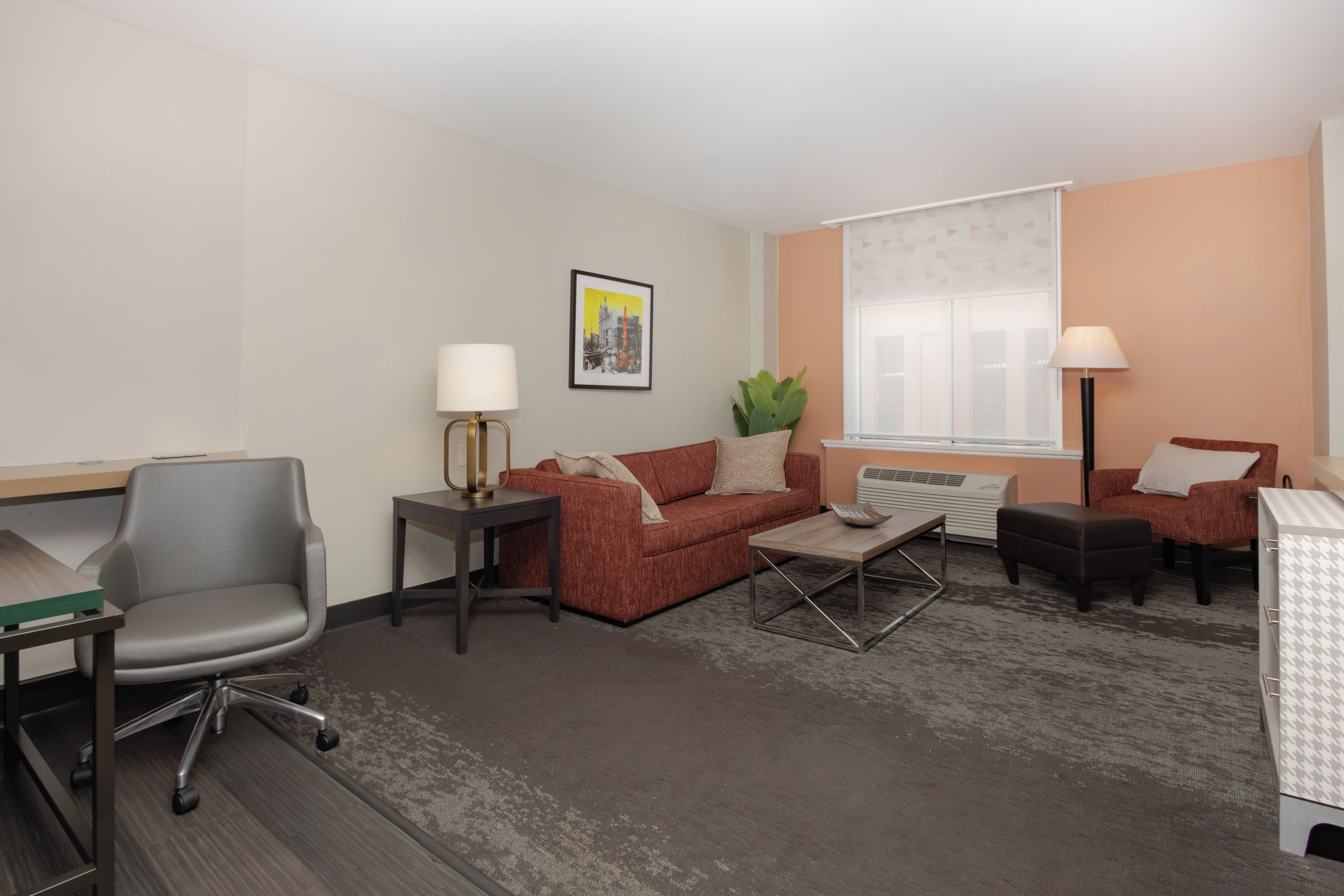A hotel room with a grey office chair, a red sofa, a small coffee table, a side table with a lamp, a potted plant, and a window with blinds.