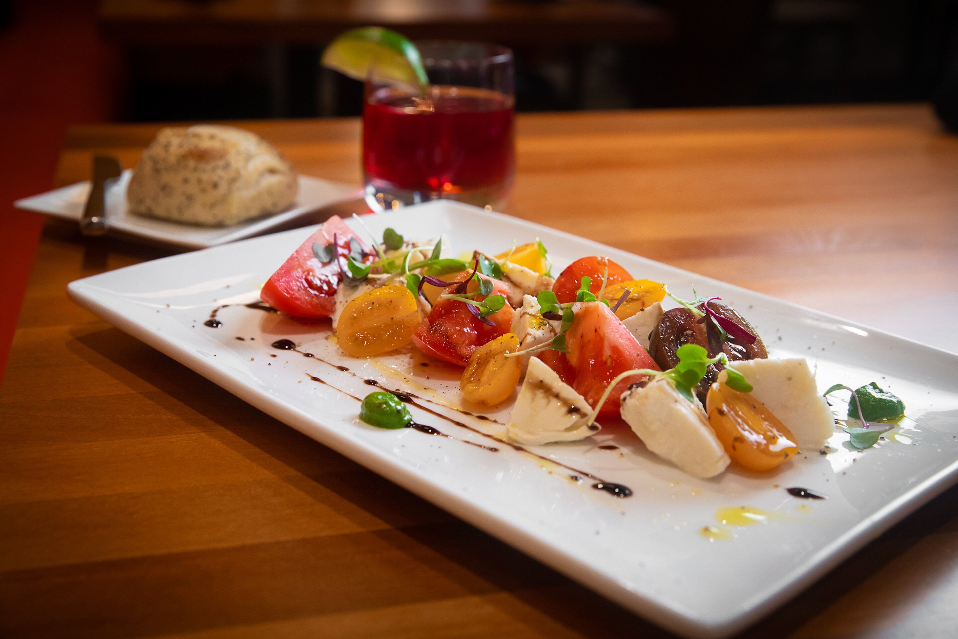 Un plato rectangular blanco con ensalada, un vaso de bebida roja y un panecillo sobre una mesa de madera.