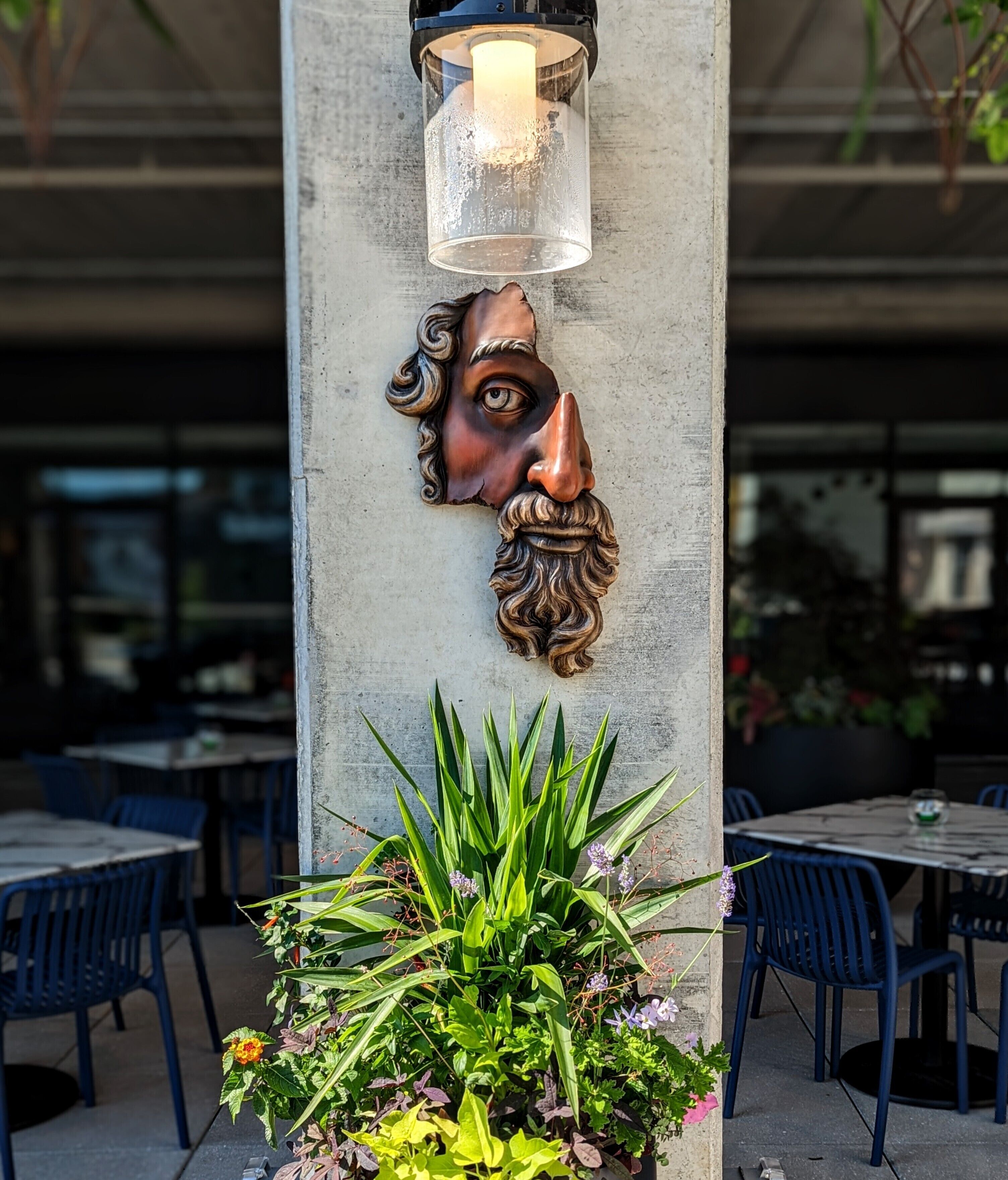 A concrete pillar with a decorative head sculpture, a hanging light fixture, and a potted plant with green foliage and purple flowers.