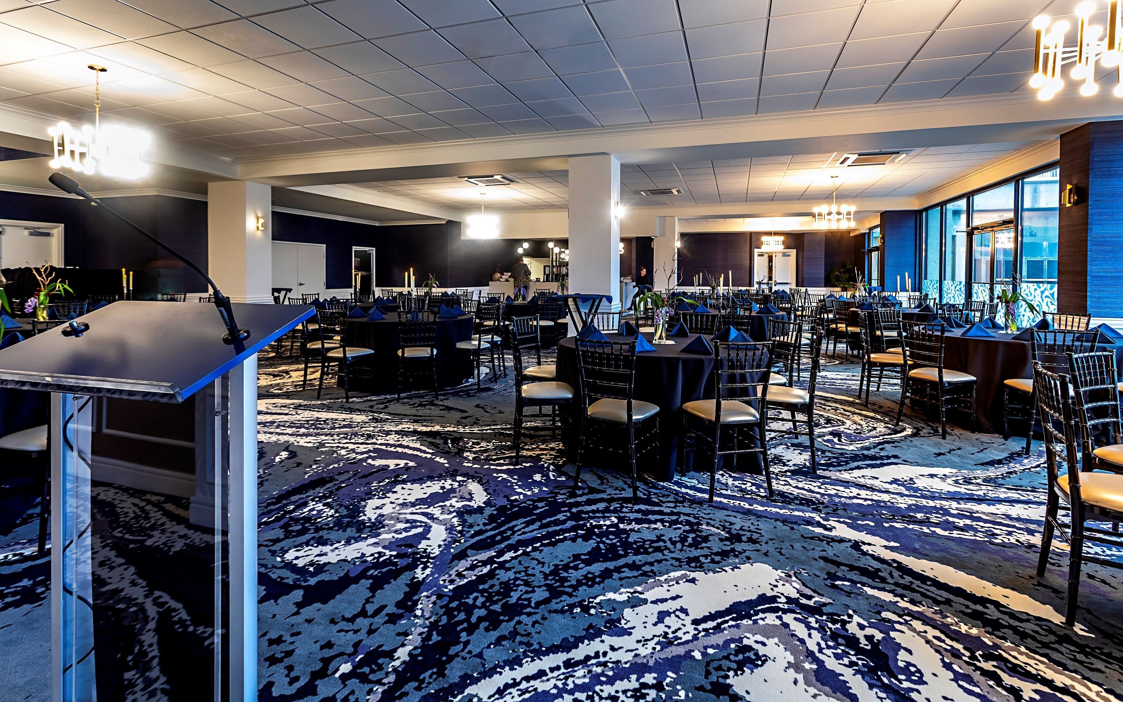 A spacious banquet hall with multiple tables and chairs, a stage with a microphone, and a patterned carpet.