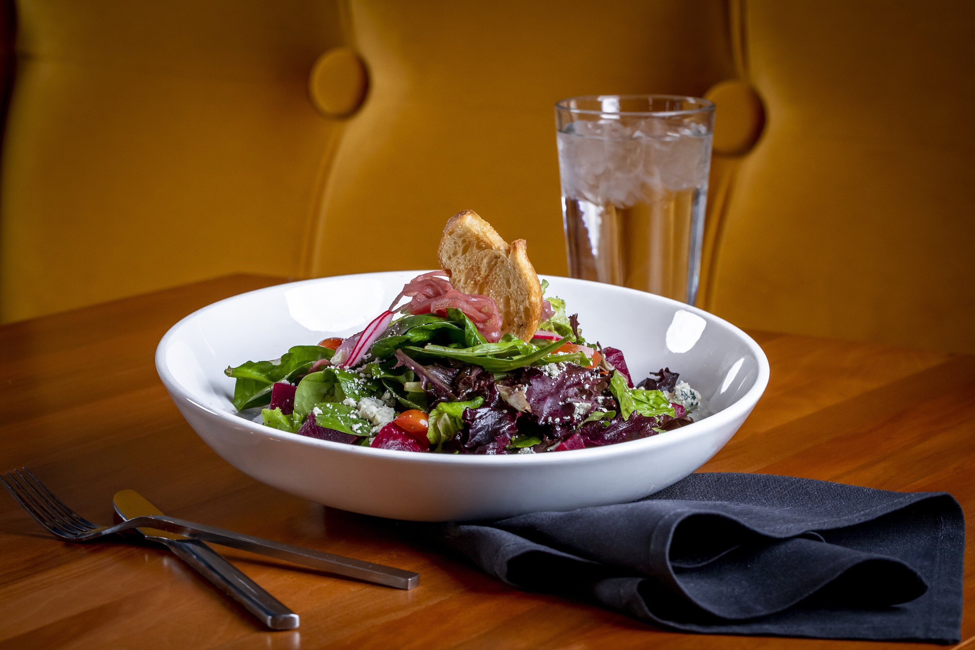 A white bowl of salad with croutons and a glass of water on a wooden table.