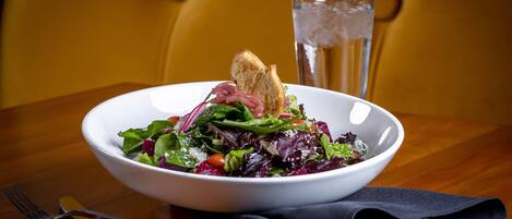 A white bowl of salad with croutons and a glass of water on a wooden table.