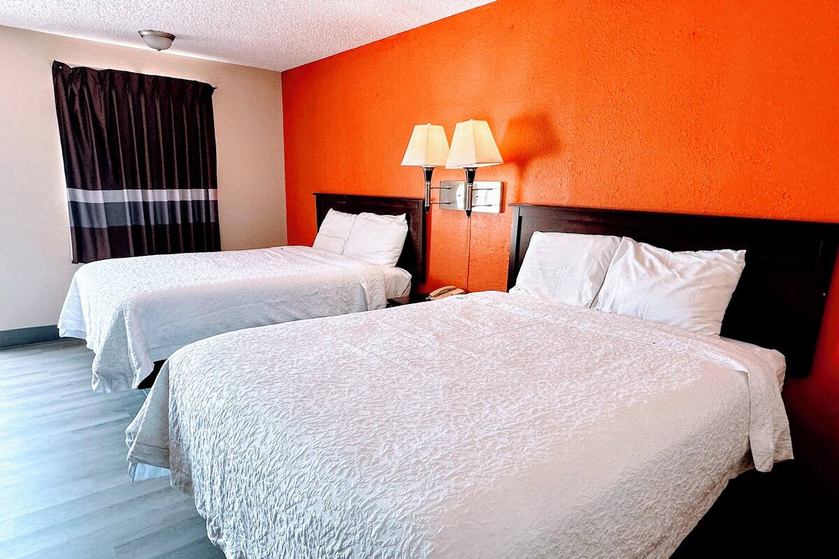 A hotel room with two beds, a nightstand, a lamp, and an orange wall.