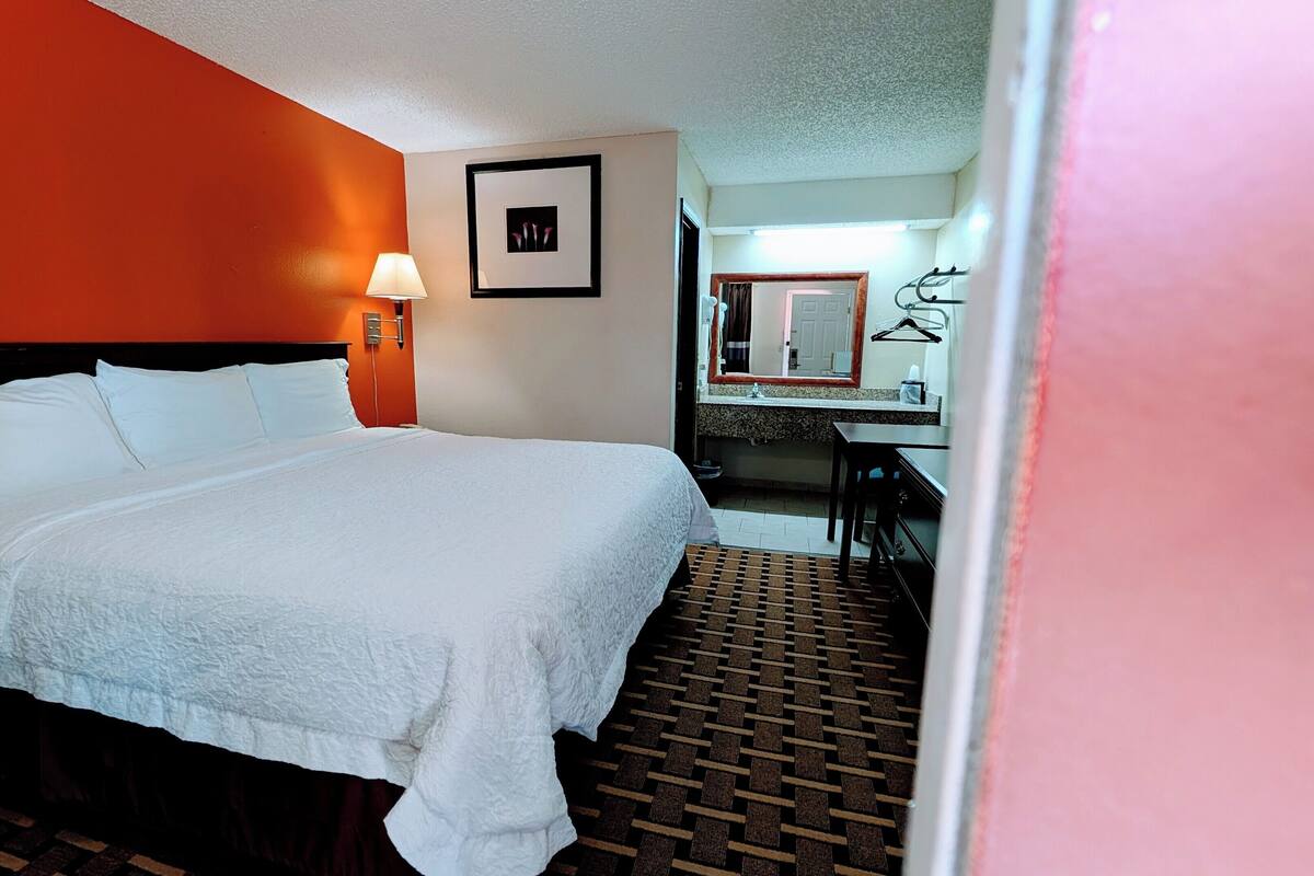 A hotel room with a large bed, a bedside lamp, a small desk with a mirror, and a bathroom visible through an open door.