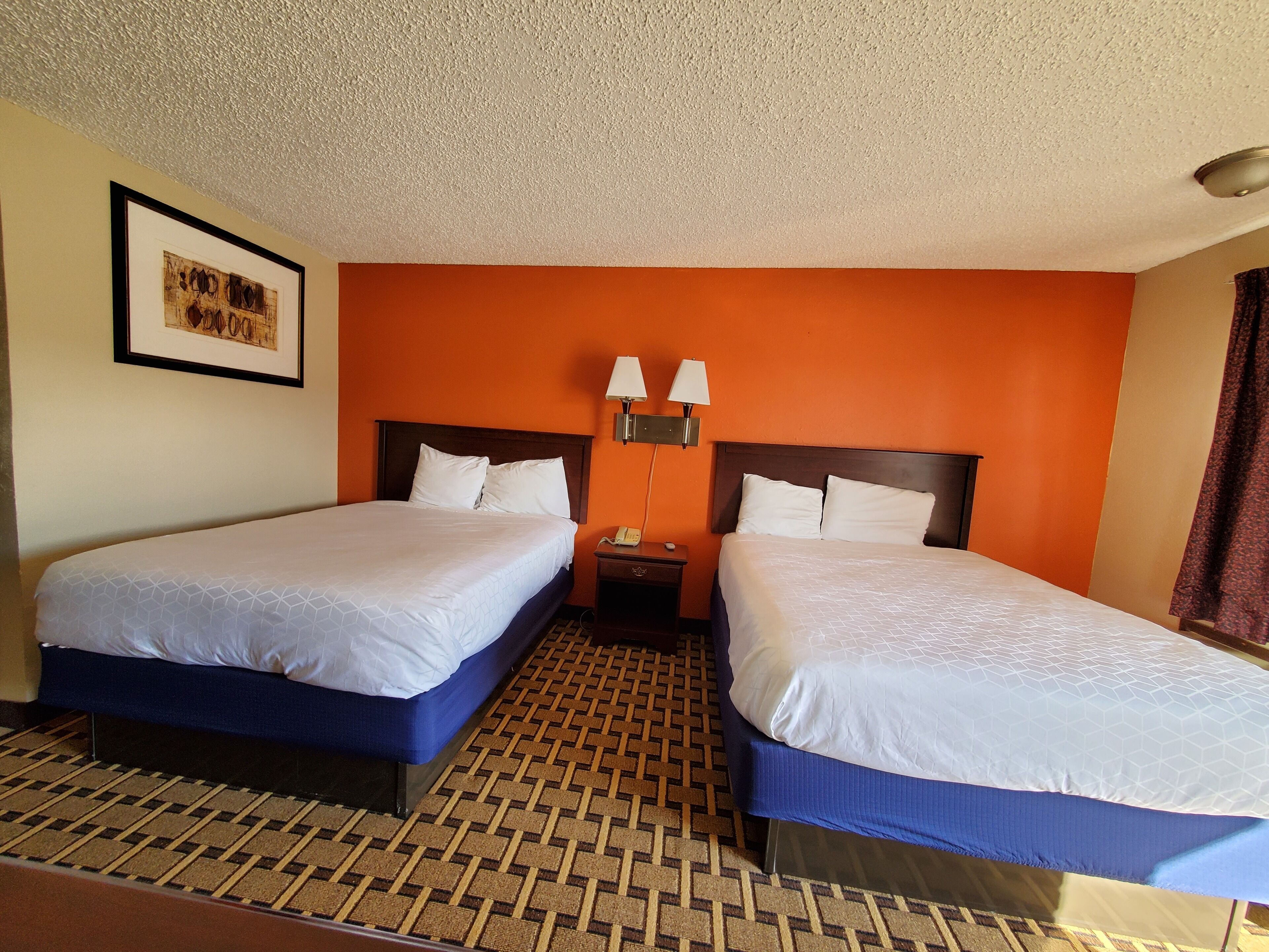A hotel room with two beds, a patterned floor, a framed picture on the wall, and a nightstand with lamps.