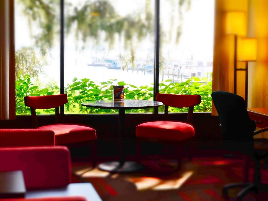 A room with a round table and four red chairs, a black chair, and a view of greenery and buildings through large windows.